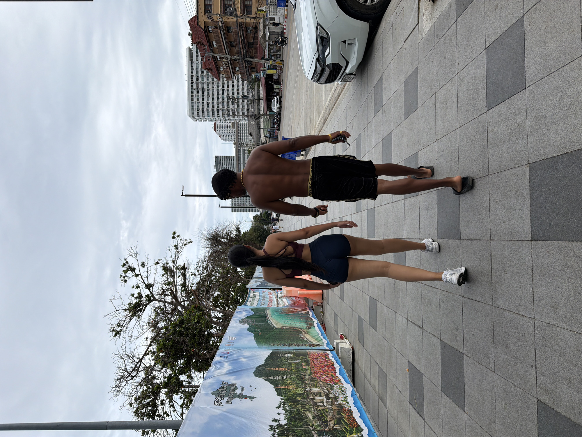 The image depicts a couple walking away from the camera on a paved sidewalk in Pattaya City, Thailand. The man is shirtless and wearing black shorts with gold trim, and a gold necklace. He is wearing black flip-flops. The woman is wearing a burgundy sports bra and dark blue shorts, and white athletic shoes. They are walking on a sidewalk with a checkered pattern of light and dark gray tiles. To their left is a long banner displaying colorful scenic images of mountains, beaches, and structures, possibly representing tourist attractions. Beyond the banner, trees are visible. To their right, a white SUV is parked, and further down the street are buildings, a lamppost, and other vehicles. The sky is overcast.