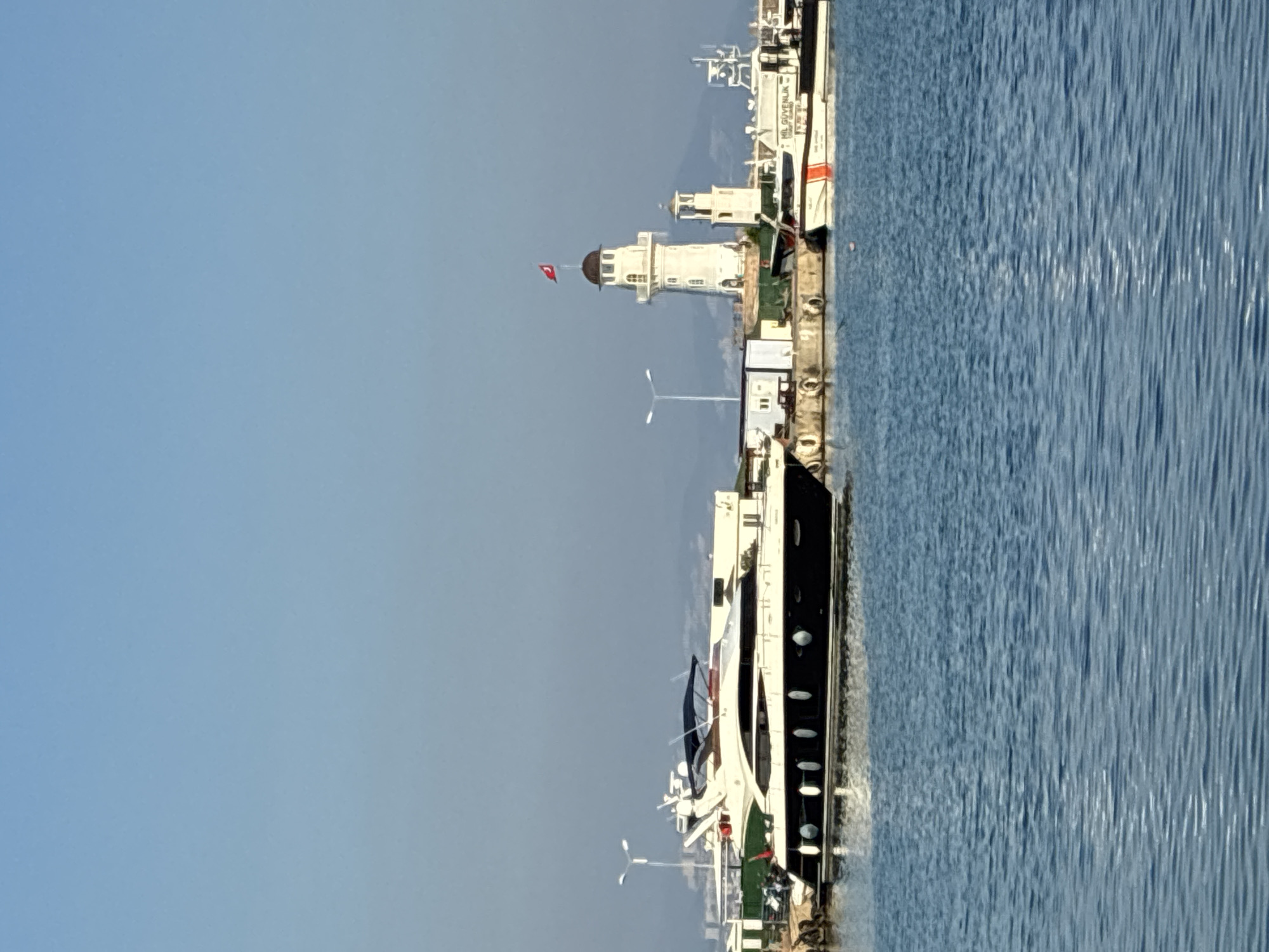 The image depicts a scenic view of the Alanya Marina in Alanya, Turkey. In the foreground, the tranquil blue waters of the harbor are visible, with gentle ripples indicating a calm day. Moored along the quay are several vessels, most notably a sleek, modern yacht with a dark hull and white superstructure, its reflection shimmering on the water. Further down the harbor, a distinctive white lighthouse stands tall, topped with a dark dome and flying a Turkish flag. Adjacent to the lighthouse, a smaller tower is also present. Several other boats, including a white and orange Coast Guard vessel, are docked. The background reveals a hazy coastline with distant buildings and mountains under a clear, pale blue sky. The scene conveys a sense of peaceful maritime activity and coastal beauty characteristic of Alanya.