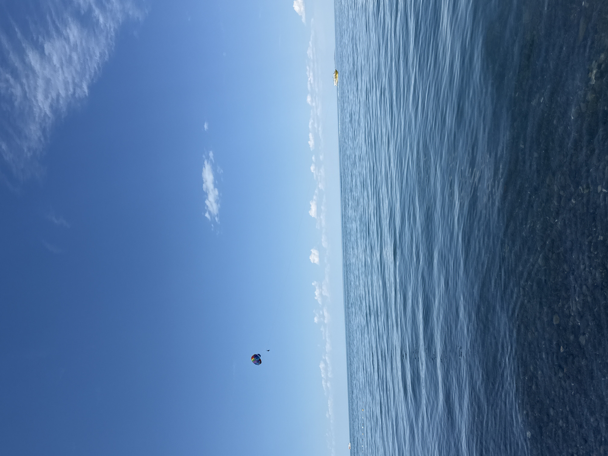 The image depicts a tranquil scene of the sea under a clear blue sky with scattered white clouds. In the distance, a colorful parasail is visible, suspended in the air with a person attached. A yellow boat is also seen on the water, moving away from the viewer. The foreground of the image shows the clear water with visible pebbles at the bottom, suggesting a shallow coastline. The overall atmosphere is serene and indicative of a pleasant day by the sea. The location is identified as Ақши, Kazakhstan.