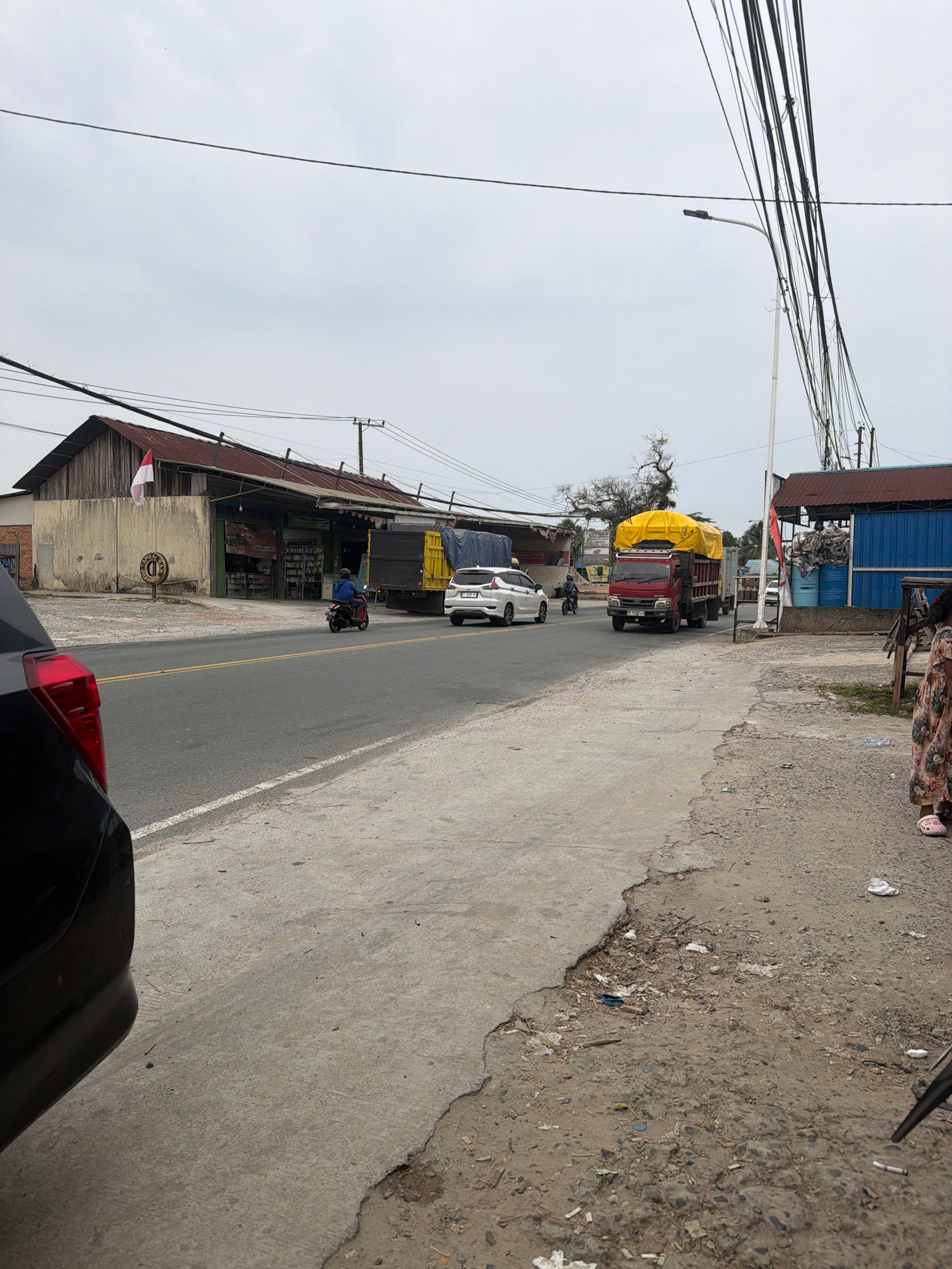 The image depicts a street scene in Balikpapan, Indonesia. In the foreground, the rear left corner of a dark-colored car is visible. The road is paved, with yellow and white lines marking the lanes. A person on a motorcycle is seen riding in the same direction as the camera. To the left of the road, there is a building with a weathered wooden facade and a corrugated metal roof. An Indonesian flag is attached to the building. Further down the road, a white car and a red truck are traveling in the opposite direction. The truck is carrying a large yellow tarp-covered load. To the right of the road, there is a blue building with a metal roof and stacks of materials. A person wearing a floral dress and pink sandals stands near this building, facing away from the camera. The environment is overcast, with power lines crisscrossing the sky. The ground on the right side of the road is unpaved and shows signs of debris.