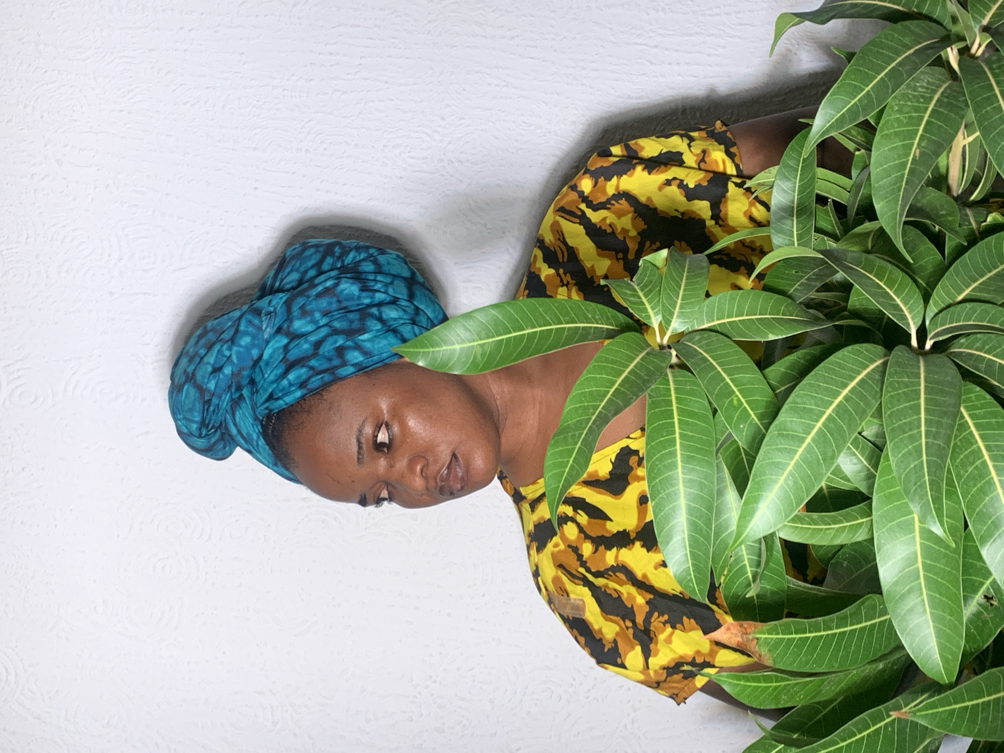The image displays a portrait of a Black woman, oriented with her head on the left side of the frame. She has a rich brown skin tone and wears a vibrant teal and blue patterned headwrap. Her top is a distinctive yellow, black, and brown pattern, resembling an abstract or animal print design. She looks downwards and slightly to her right, with a calm and somewhat contemplative expression, her lips gently parted.
The right side of the frame is dominated by a dense cluster of lush green leaves, identifiable as belonging to a mango tree due to their elongated shape and prominent venation. These leaves partially obscure her right shoulder and arm. The background behind her left side is a plain white wall with a subtle, circular textured pattern. The lighting is bright and even, suggesting it is daytime. No specific activity is depicted beyond posing for the photograph. There is no visible text in the image. The location context for this scene is Barakin Akawo, Nigeria.