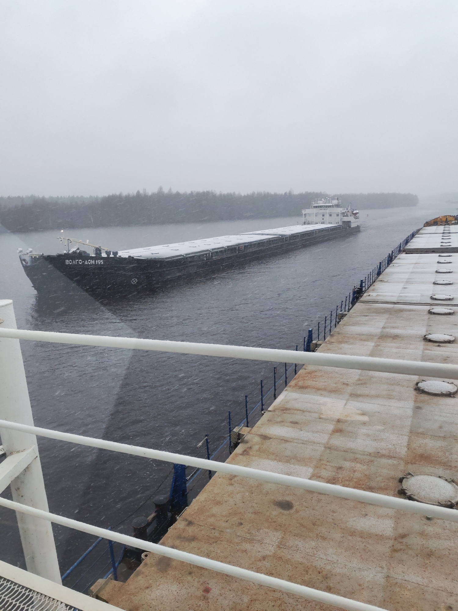 A barge, identified as "ВОЛГО-ДОН 195" in Cyrillic script, is navigating a waterway in snowy conditions. The barge is long and flat, with snow accumulating on its deck. A forested shoreline is visible in the background. The foreground shows part of a dock or pier, also covered with snow. The scene is possibly in Novokemskii, Russia, considering the barge is a Volga-Don type. There are no people visible on the barge or the dock. The primary action is the barge traveling along the waterway.