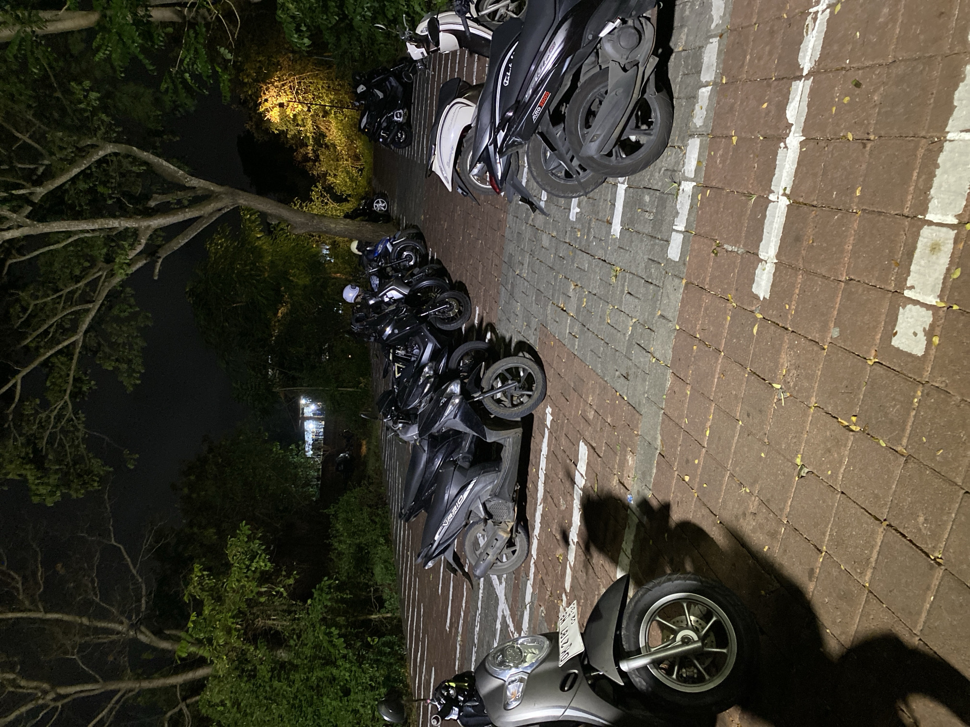 The image depicts a parking lot at night in Kuta Selatan, Indonesia, filled with many motorcycles. The ground is paved with red bricks and some gray stone tiles, with white lines marking parking spaces. Several trees with dark foliage surround the parking area, and in the background, there is a dimly lit structure with people visible inside, suggesting a restaurant or cafe. The lighting is low, creating shadows and highlighting the parked vehicles. The motorcycles are mostly dark-colored, with some white details. One scooter in the foreground is silver. The scene appears to be a typical nighttime scene in a tropical urban environment.