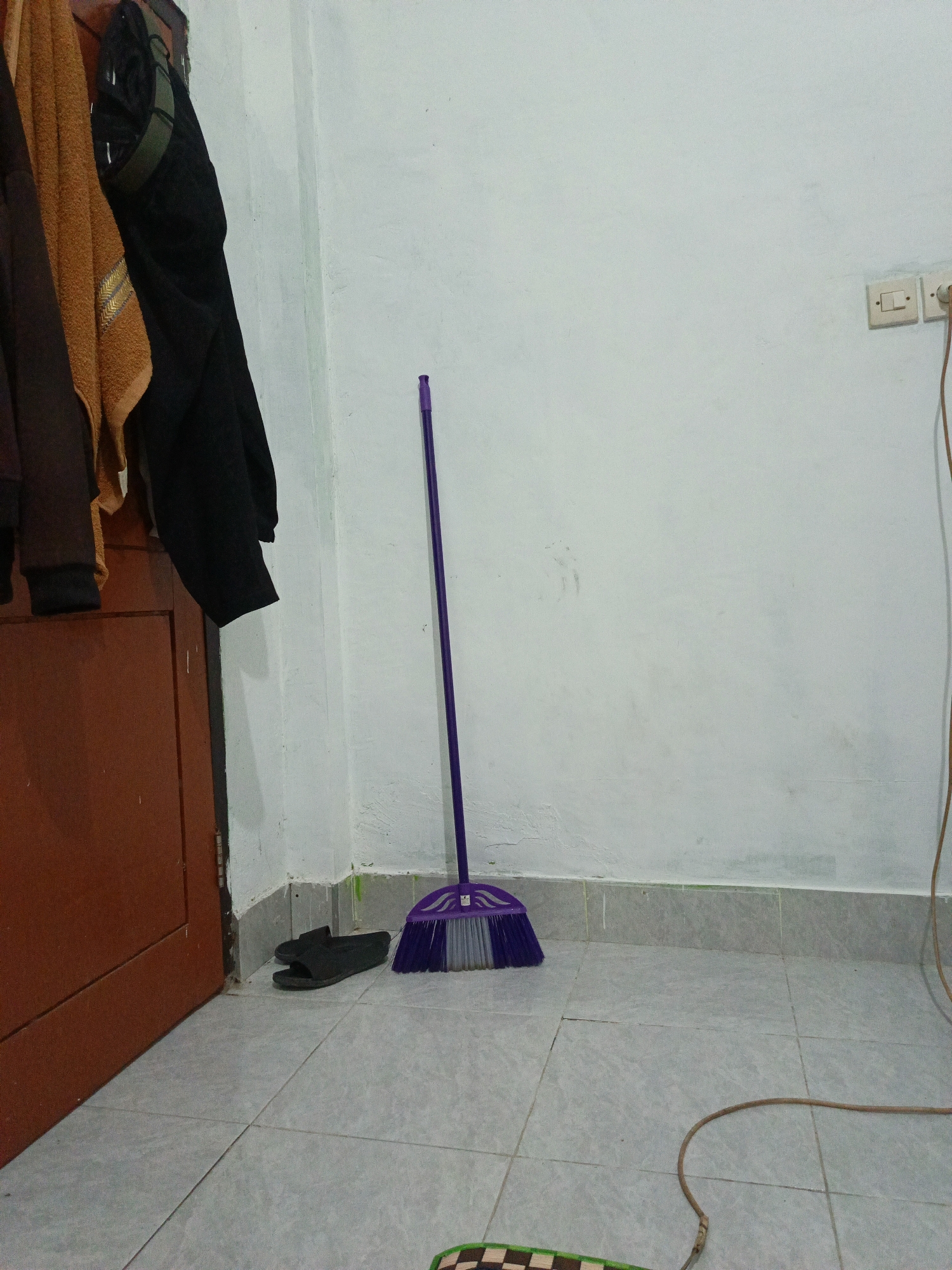 The image depicts a corner of a room with a white wall and tiled floor. To the left, a brown door is partially visible, with several items hanging on the wall above it, including a brown towel and dark clothing. In the center of the scene, a purple broom with grey bristles stands upright. To the left of the broom, a pair of black slippers rests on the floor. An electrical outlet and a light switch are mounted on the white wall in the upper right portion of the image, with a brown electrical cord extending downwards. The floor is tiled with light grey square tiles. The environment appears to be a domestic indoor space, possibly a residential dwelling in the city of Bonder, Indonesia.