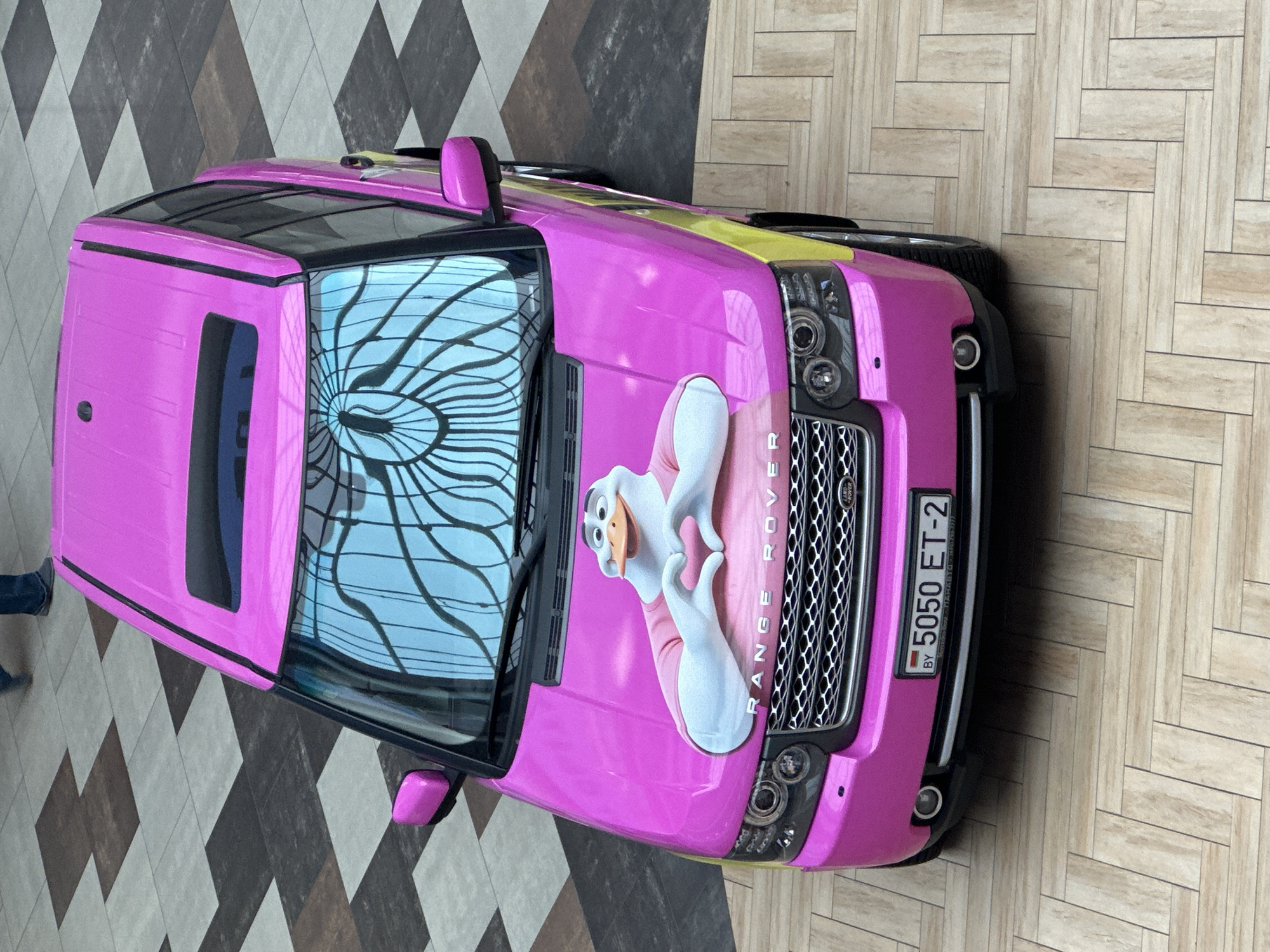 An overhead, eye-level shot captures a bright pink Range Rover parked on a checkered tiled floor with a herringbone patterned section in the foreground. The car features a cartoon duck decal on its hood, holding its wings in a heart shape, and the text "RANGE ROVER" in silver lettering. The license plate reads "BY 5050 ET-2". A person's legs, clad in dark jeans and blue shoes, are visible in the upper left corner, walking away from the vehicle. The car's windshield reflects an ornate ceiling structure. The scene appears to be in Minsk, Belarus, judging by the license plate and the architectural style of the reflected ceiling.