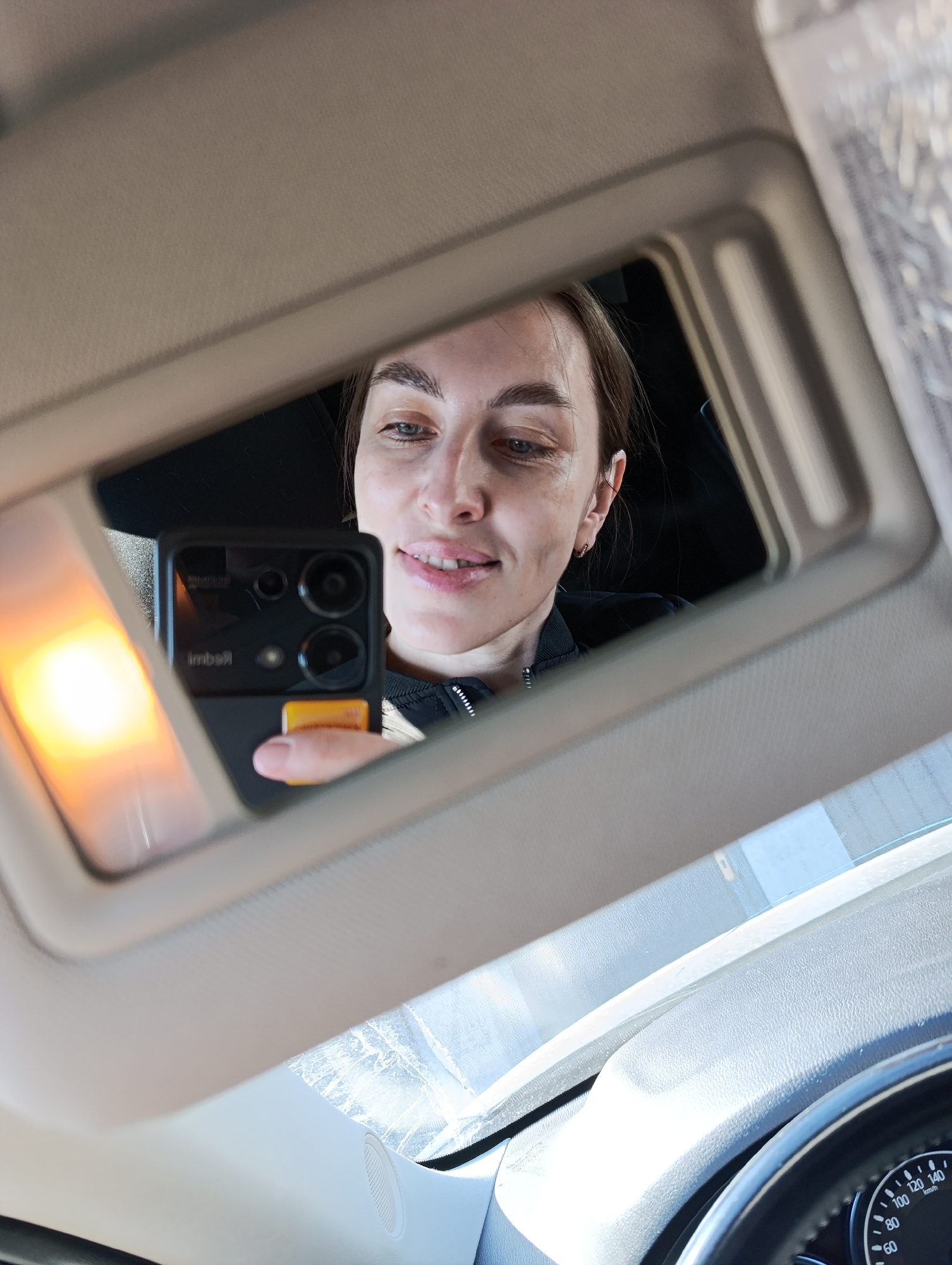 A woman is taking a selfie in the mirror of a car visor. She is holding a black smartphone with a distinctive camera arrangement. The car's dashboard and steering wheel are visible in the lower portion of the image. The location of the image is not provided in the prompt, so I cannot specify the city of Chaikovsky, Russia.