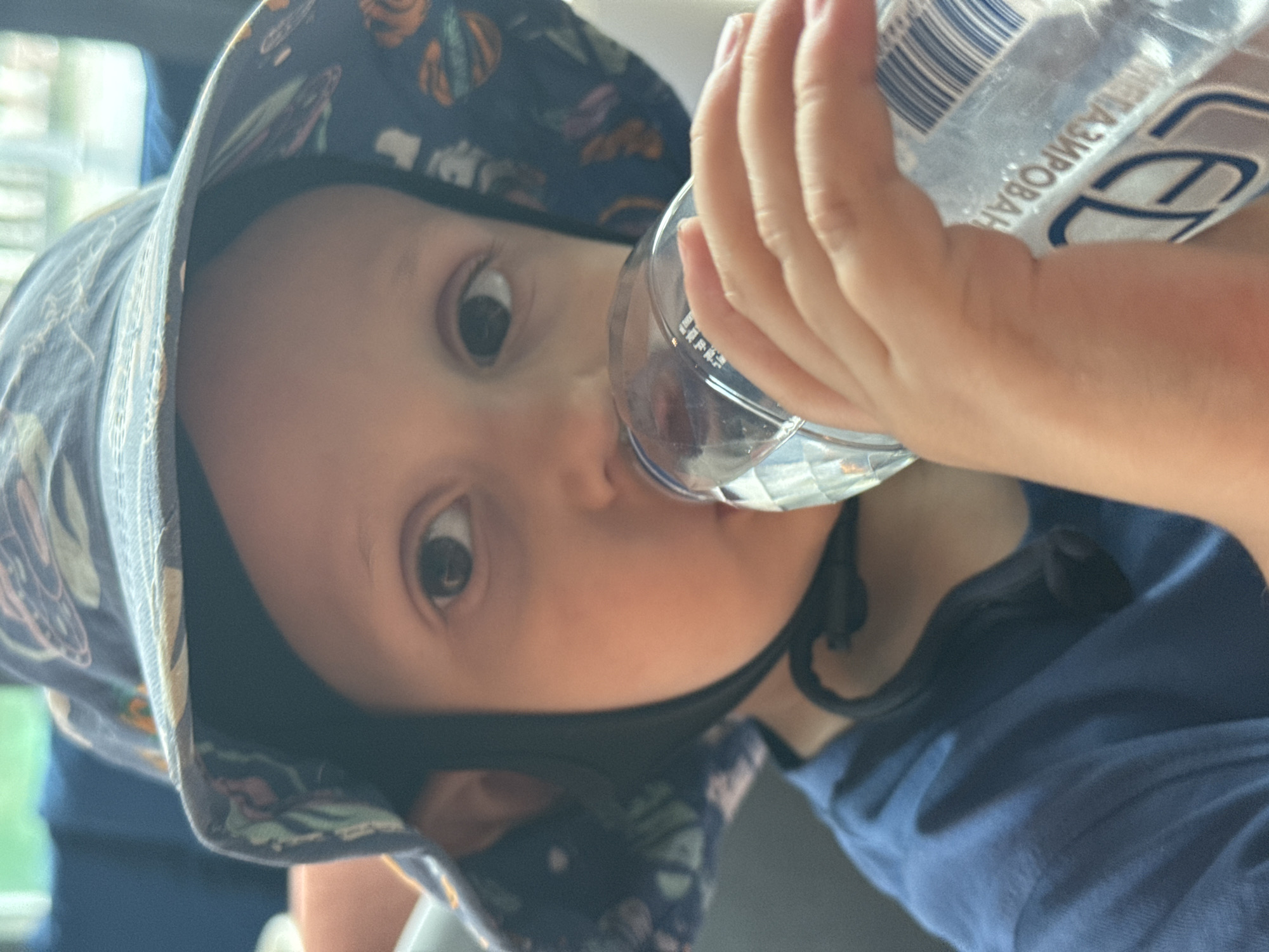 A young child wearing a blue bucket hat with a patterned brim drinks from a clear plastic water bottle held in their right hand. The child has light skin and dark eyes, and is wearing a blue t-shirt. The background is blurred, but appears to be indoors with hints of blue furniture and natural light coming from a window. Text on the water bottle is partially visible and appears to be in Cyrillic. The image is a close-up, focusing on the child's face and upper body.