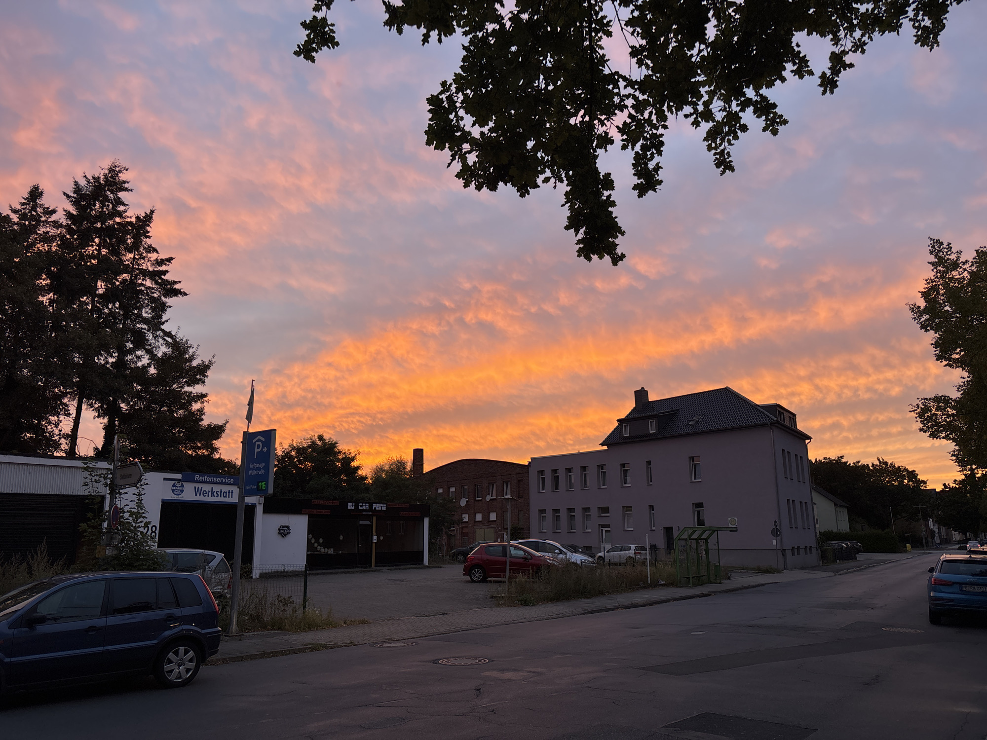 The image depicts a street scene at dusk in an urban environment. The sky is filled with vibrant orange and pink clouds, indicating a sunset. In the foreground, a dark blue station wagon is parked on the street. To the left, a building with a garage door and signage that reads "Reifenservice Werkstatt" is visible, along with a parking sign and a flag pole. Further down the street, several cars are parked in front of apartment buildings and businesses. Trees are present on the left and right sides of the frame, with some branches from an overhead tree extending into the upper portion of the image. The road is paved and shows some signs of wear. The overall scene suggests a quiet evening in a mixed-use urban area.