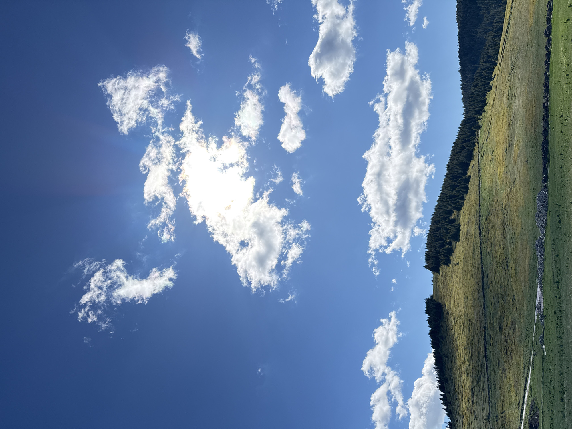 The image depicts a sunny day with a clear blue sky dotted with fluffy white clouds. The sun is partially obscured by clouds, casting rays of light downwards. In the foreground, there is a grassy hill with a scattering of trees along its crest. A dirt road winds its way up the side of the hill. The overall scene is tranquil and natural, evoking a sense of peace and serenity. The image does not contain any information about the city of Gela, Bulgaria.