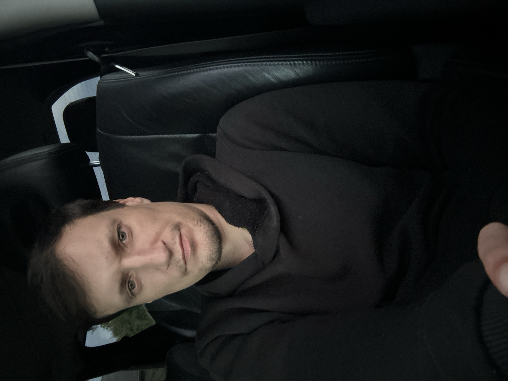 A man is sitting in the driver's seat of a car, looking at the camera. The car has black leather seats. The man is wearing a black hoodie. The background is blurred, but it appears to be daytime. There are trees and a building visible through the car window. The scene is likely in the city of Makiv, Ukraine, as indicated by the prompt.