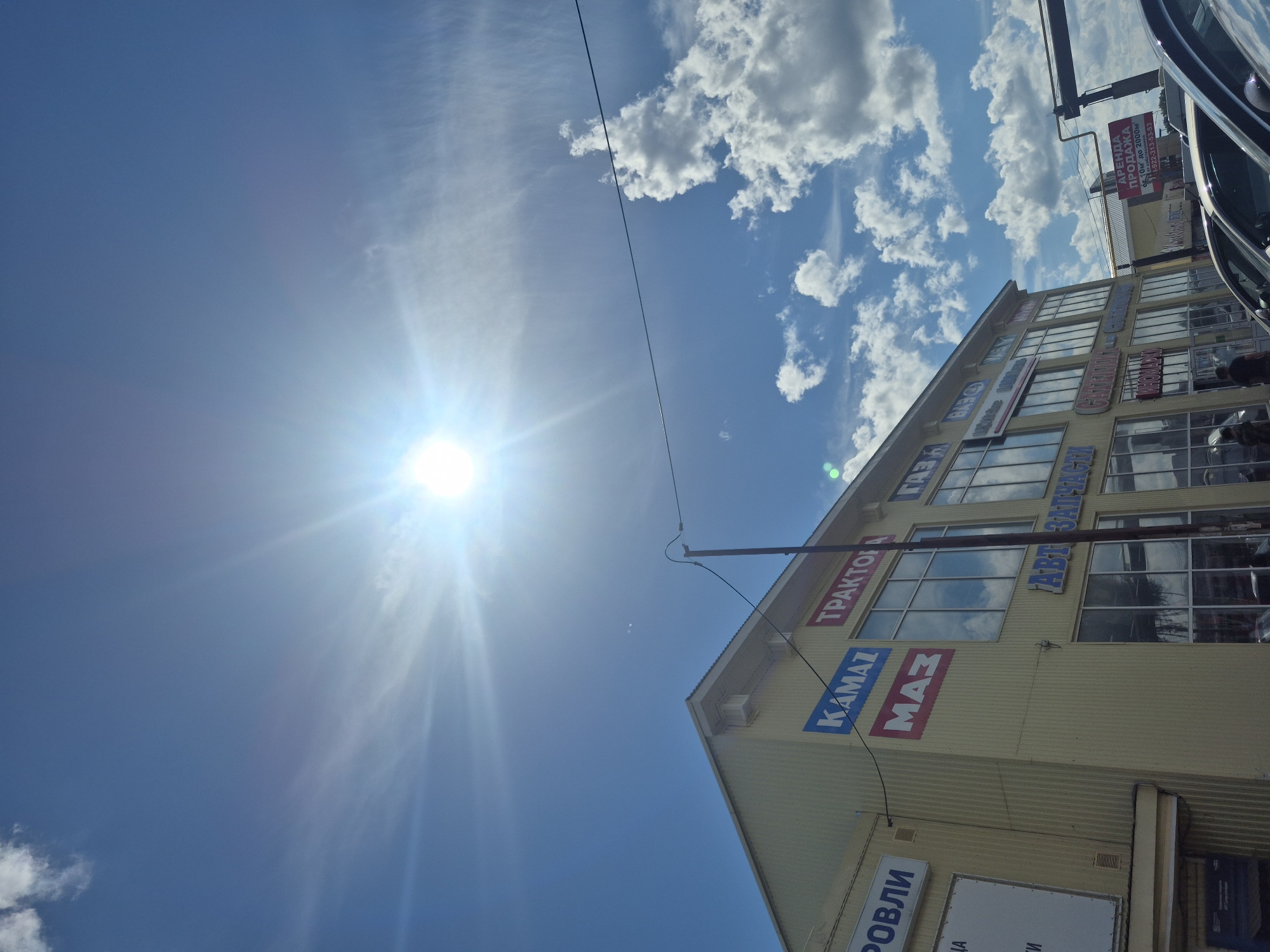 This media file captures a sun-drenched view of a commercial building in Chaikovsky, Russia. The sun is bright and prominent in the sky, with rays emanating outwards, indicating it's likely midday. Fluffy white clouds dot the clear blue sky, suggesting fair weather.
The building appears to be a multi-story structure with large windows. Several signs are visible on the facade. One prominent sign in red and white reads "АРЕНДА ПРОДАЖА" (Rent Sale), with additional details about available space and a phone number. Other signs display brand names like "KAMAZ," "MAZ," "ТРАКТОР," and "ГАЗ," suggesting the building might house businesses related to automotive parts, machinery, or retail. The overall scene depicts a commercial area on a bright, sunny day. No people are clearly discernible in the image, and the focus is on the architecture and signage of the building against the backdrop of the sky.