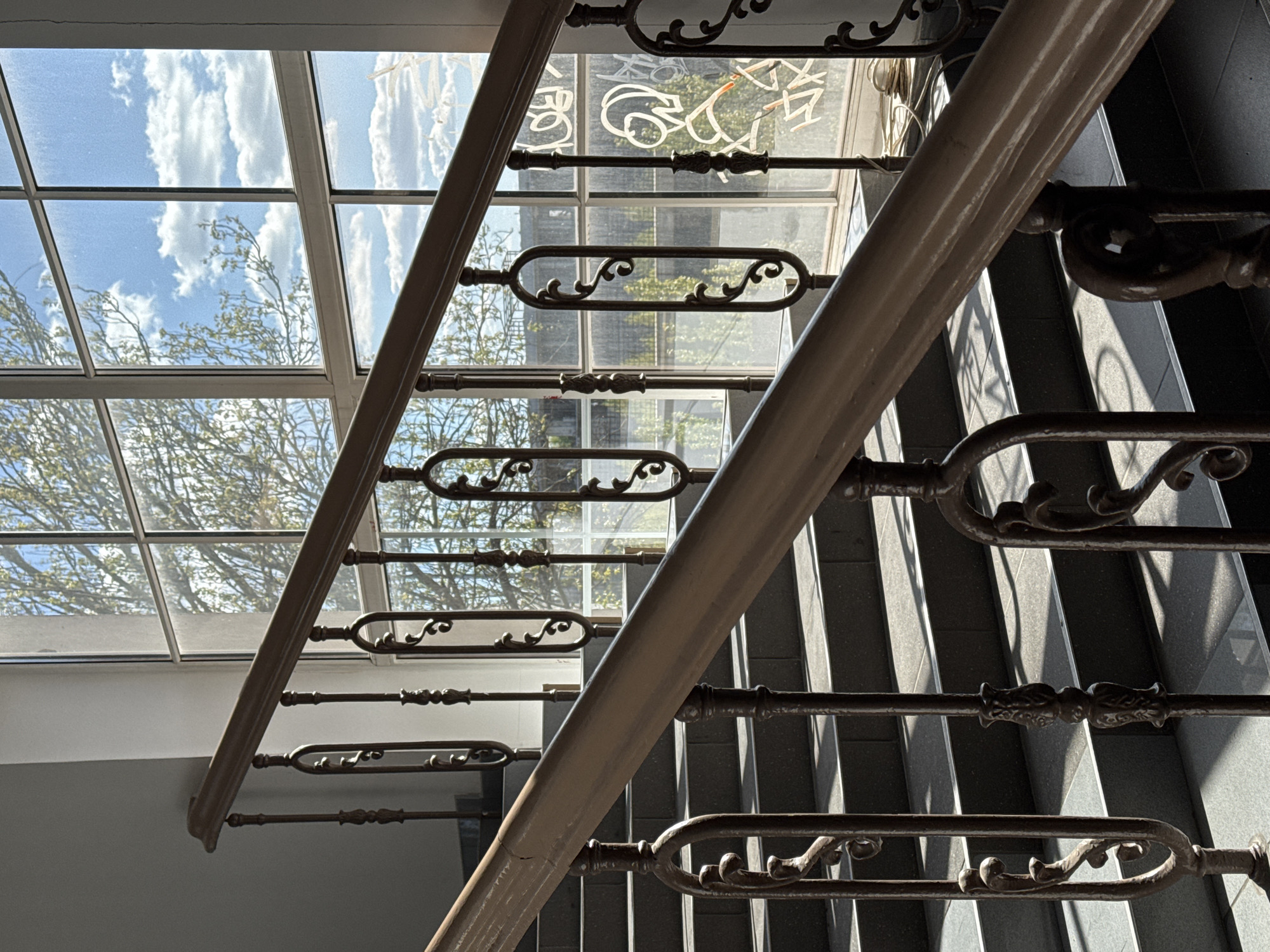 The image captures an interior staircase with ornate metal railings. Light filters through a gridded window, revealing a view of the sky with clouds and tree branches outside. The window also shows graffiti. The staircase appears to have dark-colored steps and a light-colored handrail. The location, Kryvyi Rih, Ukraine, is stated.