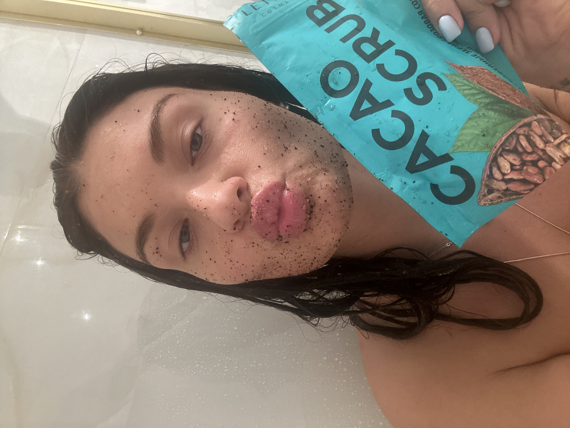 A young woman, likely in her 20s or 30s, is captured in a close-up, slightly tilted selfie, appearing to be in a bathroom or shower setting with white tiled walls and water droplets visible. Her dark hair is wet and pushed back from her face.
Her face is covered in a dark, granular scrub, particularly on her forehead, cheeks, nose, and chin, indicating she is in the process of a skincare routine. Her lips are provocatively pursed, coated in some of the scrub, while her eyes gaze towards the camera.
She holds up a light blue/teal pouch of cosmetic product labeled "CACAO SCRUB" in prominent white letters. Smaller, partially visible Russian text on the pouch includes words like "ОРОЗОВАЯ СО" and "ЗЕРНА РА," suggesting "cacao beans" or similar ingredients, which aligns with the product name. The packaging also features an illustration of cacao pods and leaves. Her fingernails, holding the pouch, are painted with light blue polish. A thin, delicate gold chain is visible around her neck. The overall scene suggests an intimate moment of personal care.