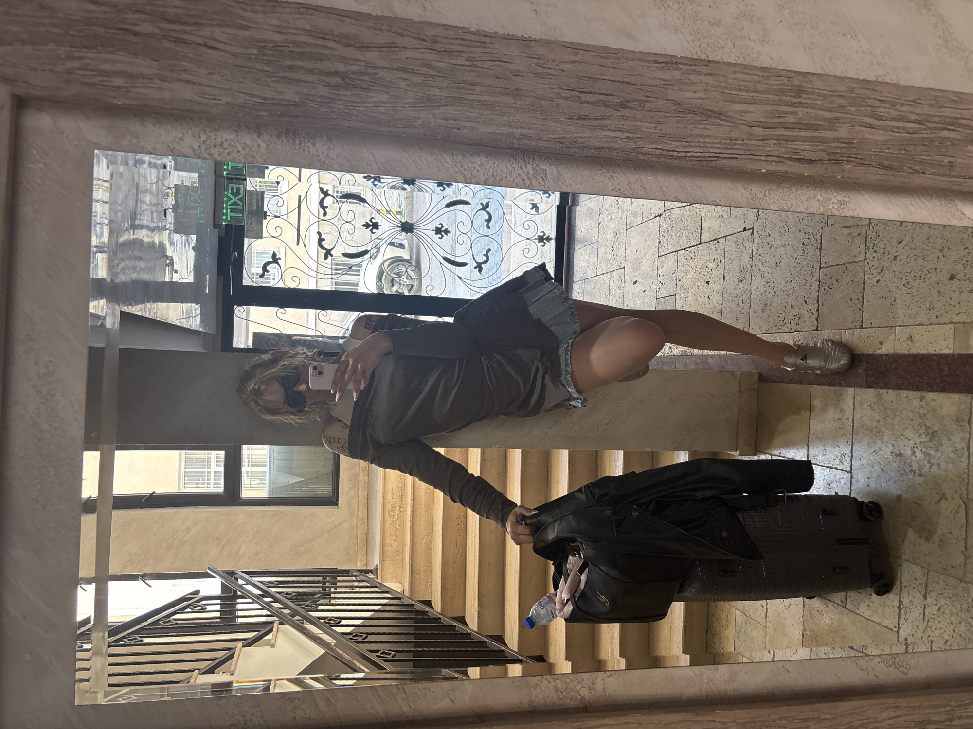 A person is captured in a mirror selfie, posing in what appears to be a building hallway or lobby in Plovdiv, Bulgaria. The image is oriented with the person lying horizontally, but should be viewed upright. The individual, with blonde or light brown hair and wearing dark sunglasses, is holding a light-colored smartphone to take the picture. They are dressed in a dark long-sleeved top and light-colored shorts or a short skirt, with a tattoo visible on their left arm. Their right foot, clad in a silver sneaker with an "Adidas" logo, rests on the tiled floor.
The person is leaning against a low, light-colored partition or banister. Next to them on the floor is a black, ridged rolling suitcase, over which a black jacket or coat is draped. A plastic water bottle with a blue cap is tucked into a pocket of the jacket or suitcase.
The mirror reflects the interior space, showing light-colored tiled or stone walls and floors, and a flight of stairs with a dark metal railing to the person's left and below. In the background, through a glass door or large window, an ornate black wrought-iron grill is visible, along with parts of an exterior building. A bright green "EXIT" sign (reflected as "TIXE") is visible near the top left of the mirror's reflection, suggesting a public or commercial building. The bright natural light indicates it is daytime. The overall setting combines modern elements (smartphone, suitcase) with more traditional architecture.