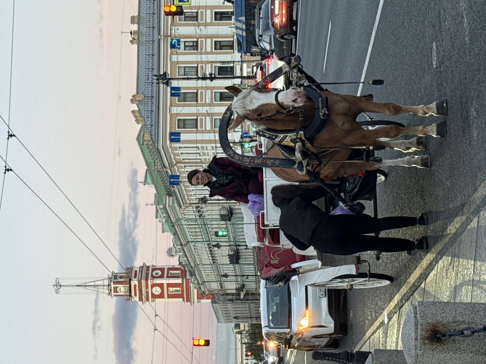 A horse-drawn carriage is on a street in Saint Petersburg, Russia. The carriage is white with red upholstery and a dark brown horse is harnessed to it. A woman in a dark jacket and pants is standing beside the carriage, attending to something within it. Another woman is seated in the carriage, looking to her left and smiling. The background shows a street with cars, traffic lights, and historic buildings with ornate architecture. A prominent red-and-white tower with a clock is visible in the distance. Overhead, power lines crisscross the sky. The time of day appears to be late afternoon or early evening, as indicated by the soft, diffused light and the colors in the sky.