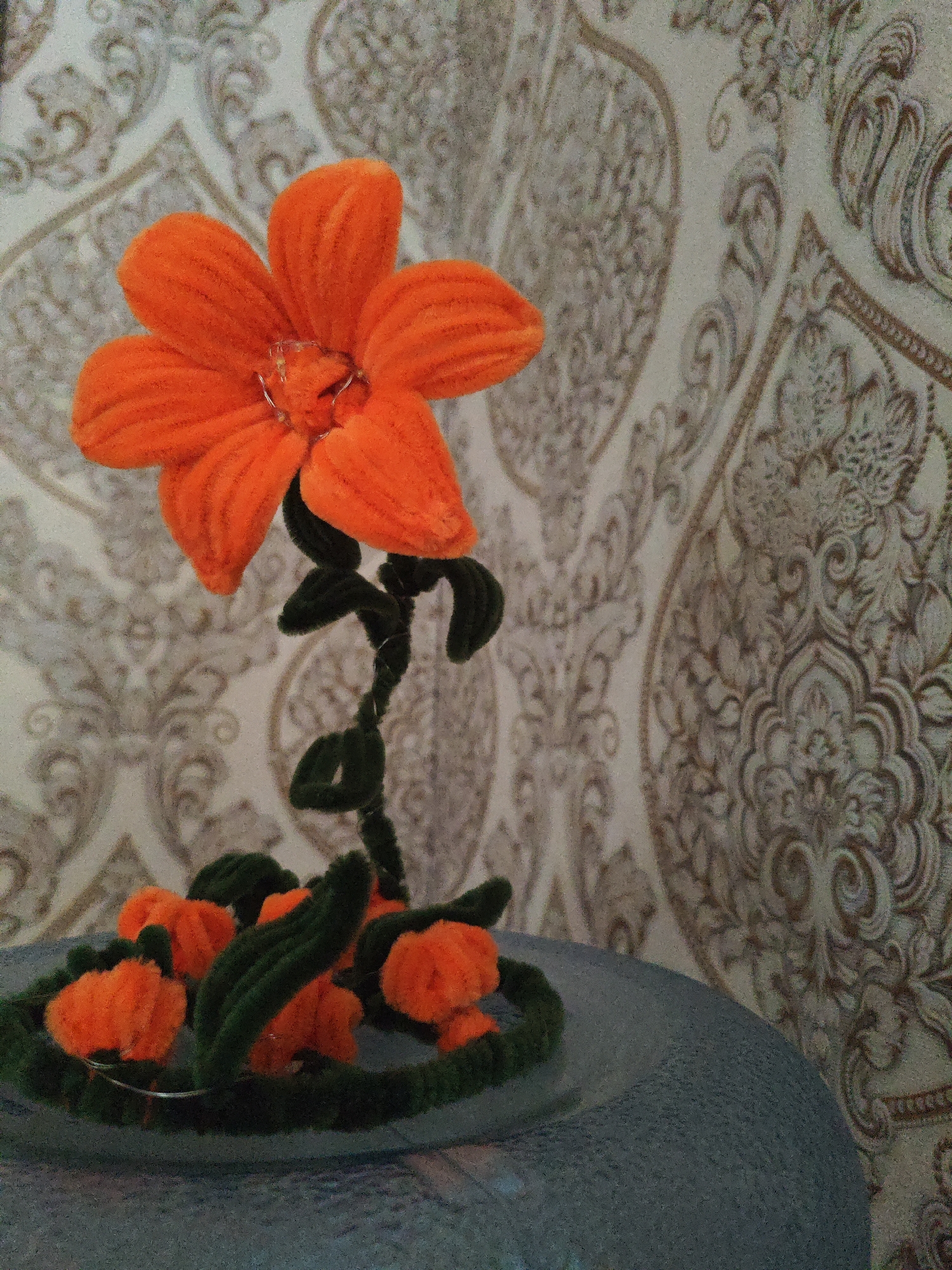 An orange pipe cleaner flower with a green stem and leaves sits on a textured, grey, circular surface. The flower is the primary subject, with five petals and a central piece. Below the main flower, several smaller orange pipe cleaner buds are arranged in a circular base with green pipe cleaner stems and leaves. The background is a light-colored wallpaper with a repeating damask pattern in muted brown tones. The lighting is somewhat dim, creating subtle shadows. The scene appears to be indoors, possibly a tabletop display. There are no people visible in the image. The location is not specified in the image itself.