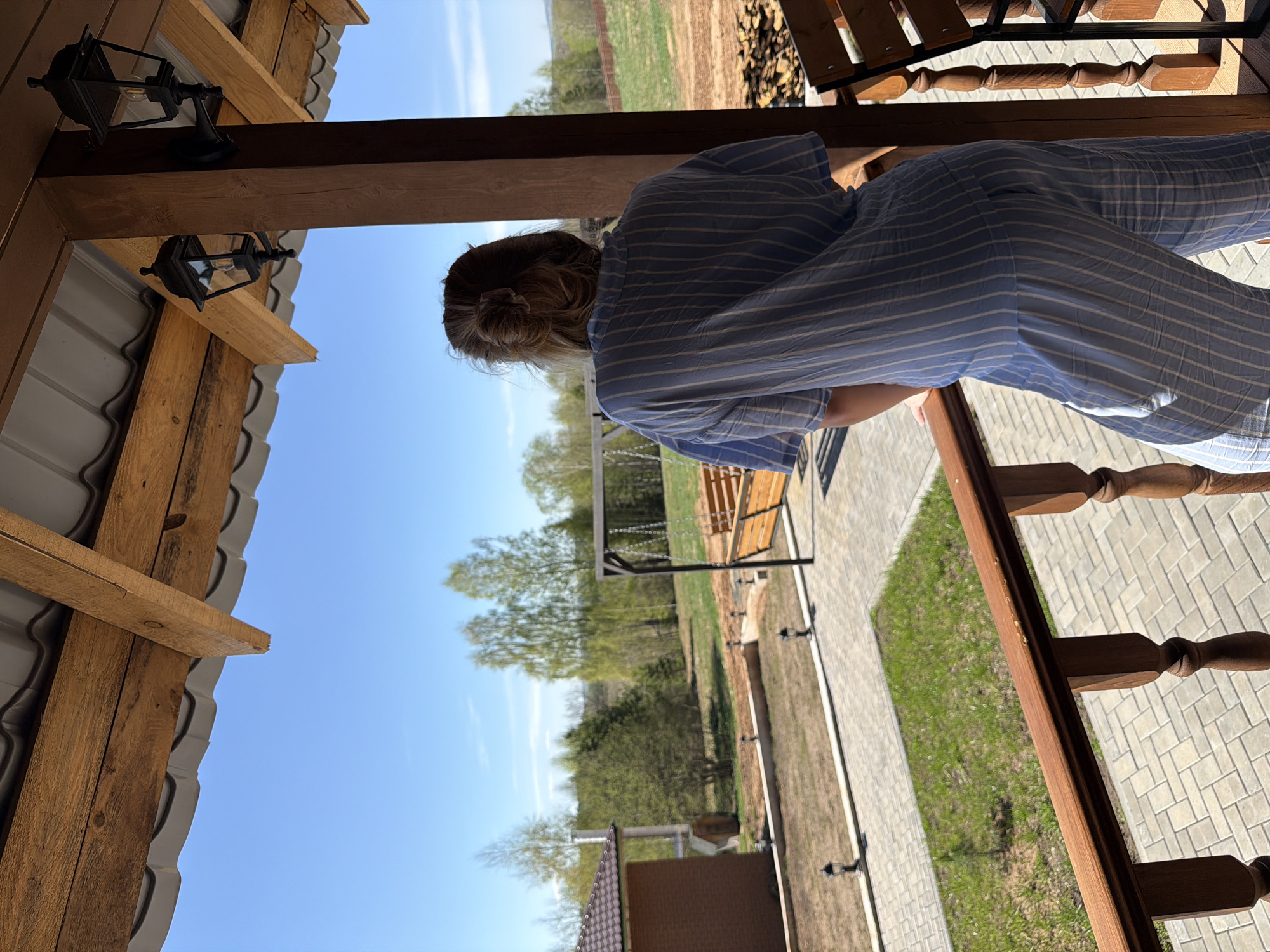 A person is standing on a porch in Kamennoye, Russia. The person is wearing a blue striped shirt and pants. The porch is made of wood and has a gray tiled floor. There is a swing set in the yard. The sky is blue and there are some trees in the background.