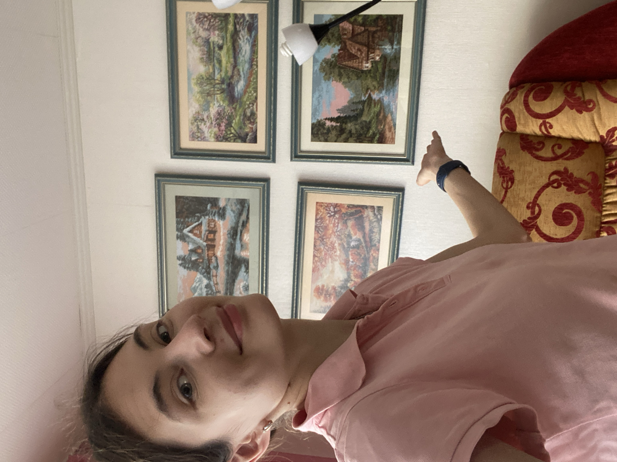 The image depicts a young woman in a pink polo shirt, standing in a room with four framed landscape paintings on the wall. The paintings appear to be idyllic rural scenes, one showing a snow-covered cottage, another a stream with blooming trees, a third a forest with autumn foliage, and the fourth a waterside cottage at dusk. The woman is smiling and looking towards the camera, with her left arm extended, as if pointing or gesturing. She is wearing a dark blue wristband. Behind her, a portion of an armchair with a red and gold patterned fabric is visible. The overall impression is a casual indoor portrait. This image was taken in the city of Votkinsk, Russia.