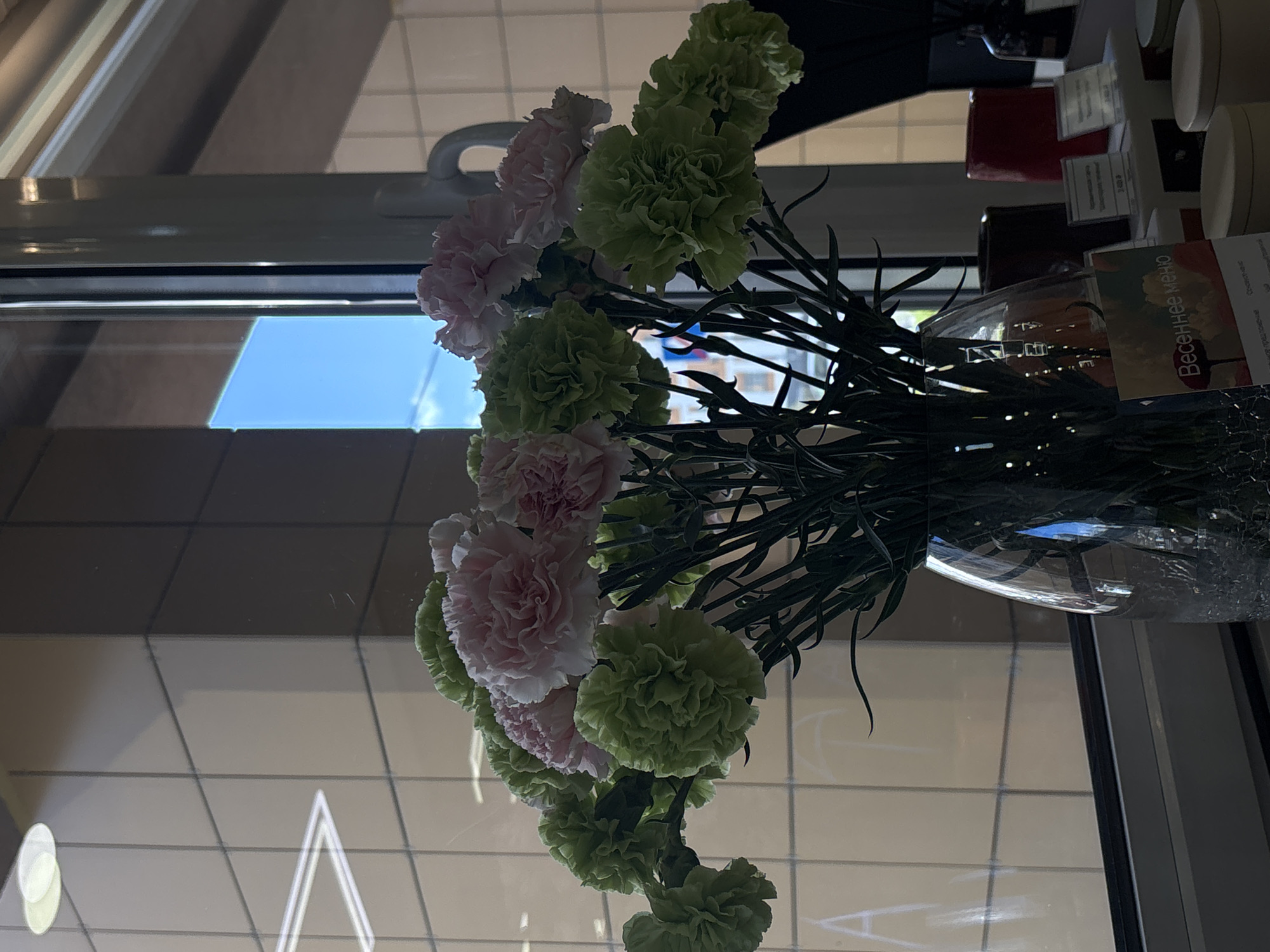The image displays a vibrant bouquet of carnations in a clear glass vase, positioned indoors next to a large window. The flowers are a mix of soft pink and pale green, characterized by their dense, ruffled petals and long, dark green stems.
Through the window, a bright blue sky is reflected or visible, suggesting clear daytime conditions. The window glass also shows reflections of geometric patterns, possibly from external buildings or interior structures. The vase, partially filled with water, holds the bouquet upright.
To the right of the vase, on a surface behind the flowers, several objects are visible, including dark red and black cylindrical containers, as well as white, possibly ceramic, cylindrical items. A small card or sign is also present, bearing the Cyrillic text "Весеннее меню," which translates to "Spring menu," indicating a potential commercial setting like a cafe or restaurant. The floor or surface below the window features a light-colored, tiled pattern.
The scene is brightly illuminated by natural light, enhancing the fresh appearance of the flowers. No people are visible.