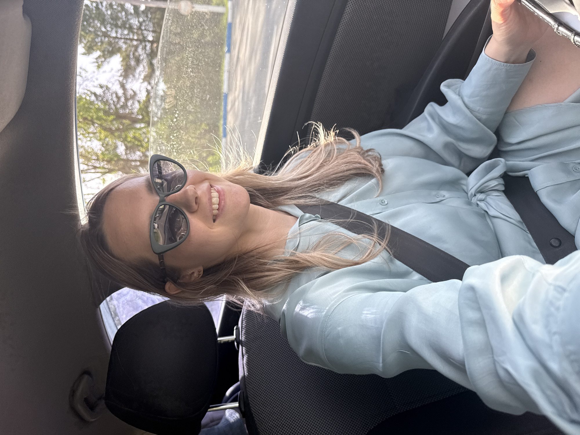 A woman with long blonde hair is sitting in the driver's seat of a car, wearing a light blue button-down shirt and sunglasses. She is smiling and wearing a seatbelt. The car is parked, and through the window, trees and a blurred urban landscape are visible. The location is potentially in Chaikovsky, Russia, based on the prompt, but there are no visual cues to confirm this definitively.