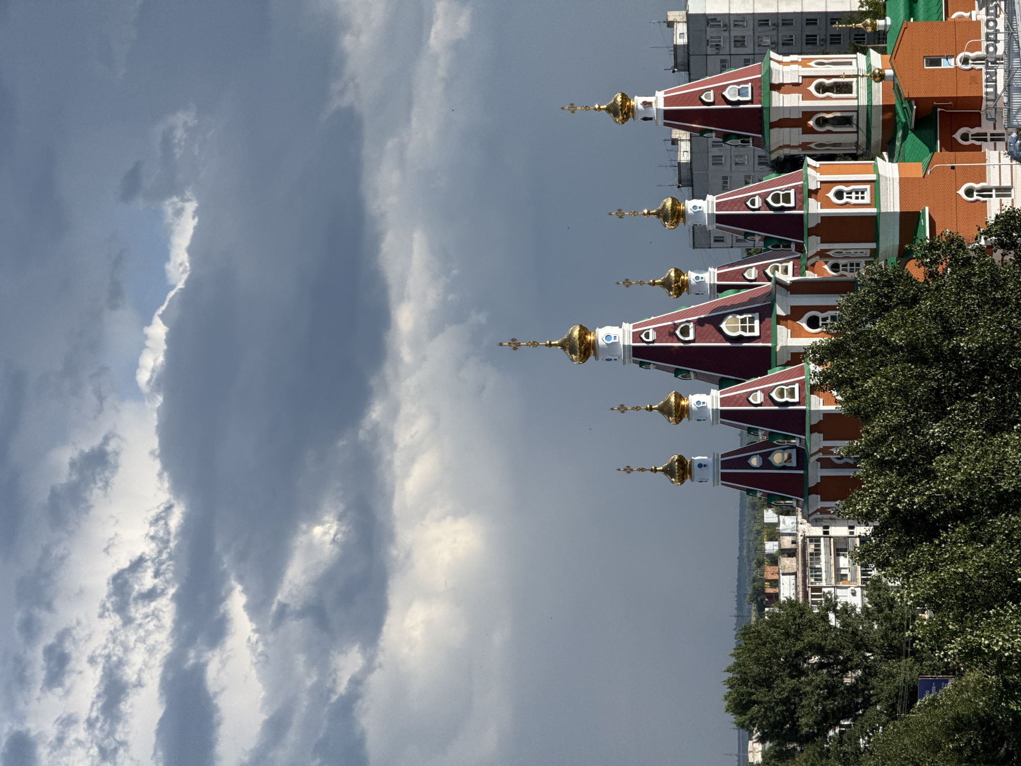 This media file, taken in Otradny, Russia, depicts a striking architectural ensemble of several domed buildings, likely a church or monastery complex, set against a dramatic sky. The buildings feature red tiled roofs, green accents, and prominent golden domes topped with crosses, indicative of Russian Orthodox architecture. The foreground is dominated by a dense green tree on the right side of the frame. In the background, a tall, modern apartment building with visible windows and balconies stands in contrast to the older religious structures. The sky is filled with a mix of dark, heavy clouds and brighter, illuminated areas, suggesting a day with dynamic weather.
The composition is unusual due to its vertical orientation, making it appear as if the buildings are either rising from the clouds or falling from the sky. This orientation, combined with the intense sky, creates a somewhat surreal and awe-inspiring visual. The time of day is not precisely discernible but appears to be daytime, possibly late afternoon or early evening given the lighting and cloud formation. No people are visible in the frame.
There is visible text in the upper right corner of the image, appearing as white lettering on a blue background. The text is partially obscured and difficult to read definitively, but it seems to be Cyrillic.
The scene captures a blend of historical and contemporary architecture within a potentially atmospheric weather condition, evoking a sense of grandeur and perhaps a touch of the sublime.