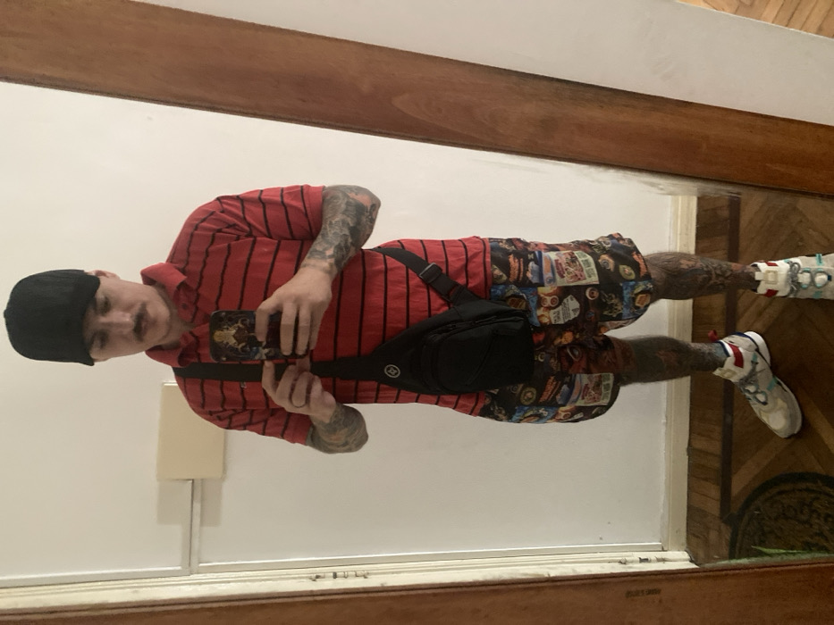 The image depicts a person taking a mirror selfie indoors. The image itself is rotated 90 degrees clockwise, showing the person standing vertically in the reflection.
The subject is an adult, appearing to be male, wearing a casual outfit. He has a black cap or beanie on his head and a mustache. Both his arms and legs are extensively covered in tattoos, visible through his short sleeves and shorts.
He is wearing a red short-sleeved polo shirt with thin black horizontal stripes. His shorts are multi-colored and feature a busy pattern with various patches, possibly depicting cartoons, comic book panels, or retro imagery. A black fanny pack or sling bag with a small, circular logo is worn diagonally across his chest and torso. He is holding a smartphone with a distinctive, possibly gold or ornate, case in his right hand, positioned as if taking the photo. A ring is visible on his left hand. His footwear consists of light-colored athletic shoes (sneakers) with colorful accents (red, blue, white, green) and matching or patterned socks.
The background reflects a simple indoor setting. There are white walls, and a wooden beam or door frame runs horizontally across the top and bottom of the frame (due to the reflection and rotation). A wooden floor, possibly parquet, is visible at the very bottom (reflected). Behind the person's left arm, a rectangular white object, possibly an intercom or thermostat, is mounted on the wall. No discernible text is visible in the image.