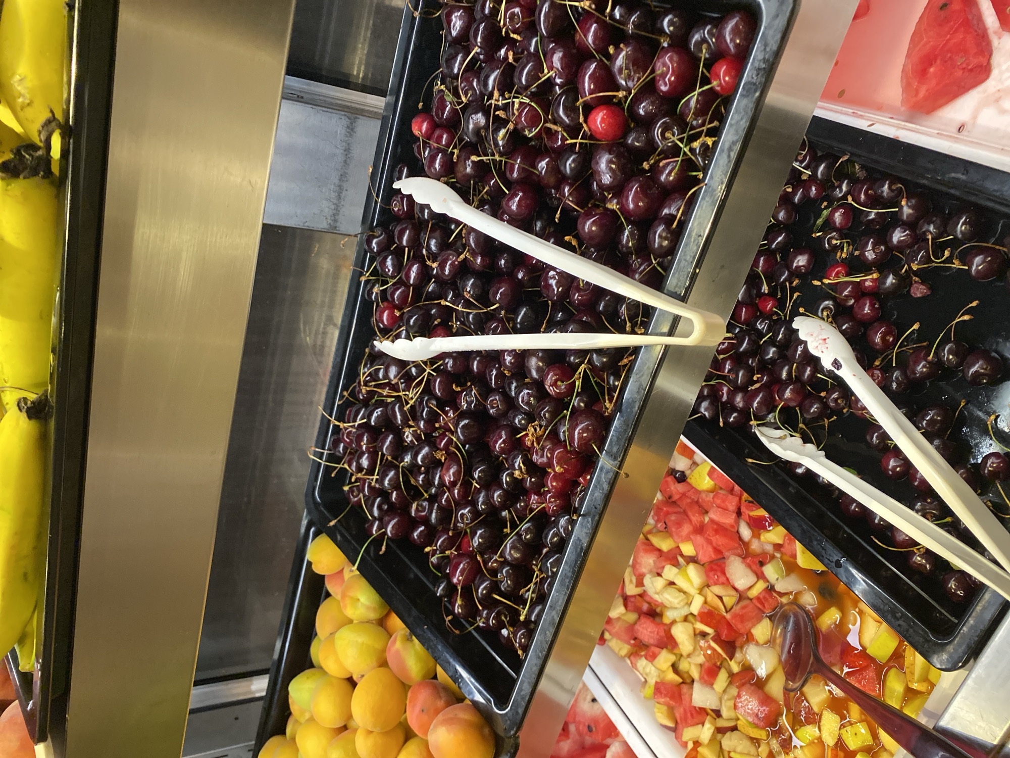In a retail environment, likely a grocery store or market in Aksu, Turkey, a display of fresh fruit is presented. The foreground is dominated by two black plastic trays filled with dark red cherries. White plastic tongs rest in the upper tray. To the left of the cherries, a white rectangular container holds a fruit salad composed of diced watermelon, apples, and what appear to be peaches or apricots, with some liquid at the bottom, and a dark red spoon partially submerged. Above the cherries, another black plastic tray contains a pile of apricots. In the background, a shelf displays a bunch of ripe yellow bananas. The scene is brightly lit, suggesting an indoor commercial setting.