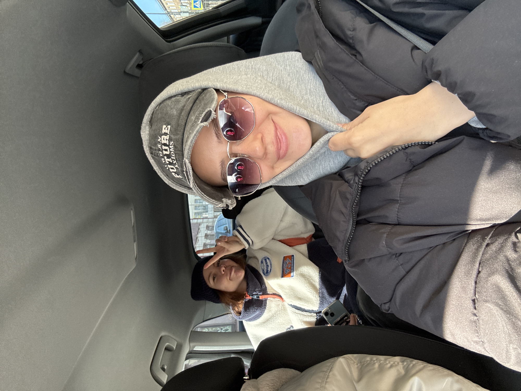 Two women are sitting inside a car. The woman in the front is wearing a grey hooded sweatshirt, sunglasses, and a black jacket. The woman in the back is wearing a black beanie and a white fleece jacket. The photo appears to be taken inside a vehicle. Based on the visible signage in the background, the location is Krasnoyarsk, Russia.