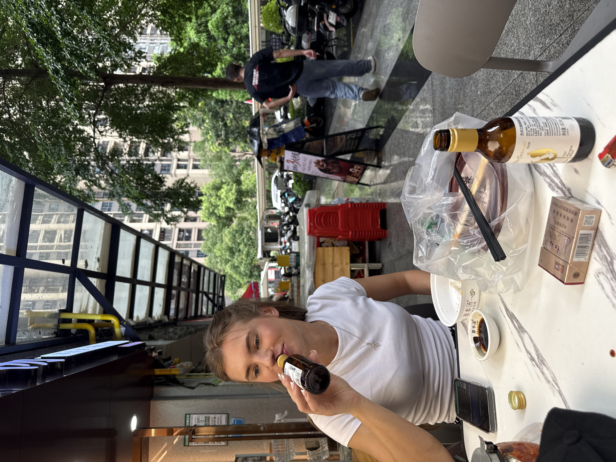 In Haizhu District, China, a woman in a white t-shirt and a silver necklace is sitting at a marble-topped table outdoors. She is holding a small brown bottle to her nose, appearing to smell it. On the table, there are other items including a white bowl with dark liquid, chopsticks, a plastic bag, a cigarette pack, and a mobile phone. In the background, there are trees, buildings, and a street scene with parked motorcycles and a man in a black t-shirt and jeans walking away. The image captures a casual moment of a woman enjoying an outdoor meal or break, with various objects suggesting a common urban setting. The time of day appears to be daytime, given the natural light. Visible text includes "草本汤" (Herbal Soup), "方风味" (Flavor), "100", and warnings about smoking.
