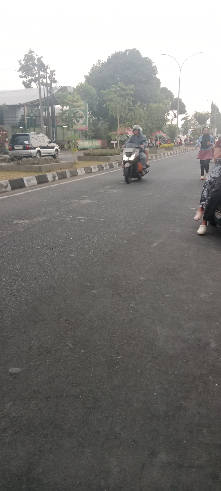 In Ngemplak, Indonesia, a street scene unfolds with a motorcycle rider in the foreground, wearing a blue jacket and a helmet, traveling on a paved road. To the left, a dark SUV is parked near a building with a white awning and signage in green and white. In the background, lush green trees dominate the landscape, with a cloudy sky overhead. Several people are visible on the right side of the road, with one individual in a light blue shirt and pink skirt standing, while others are seated. The road has a visible median with concrete dividers and black and white stripes. The overall atmosphere is one of a typical day on a moderately busy street in a town.