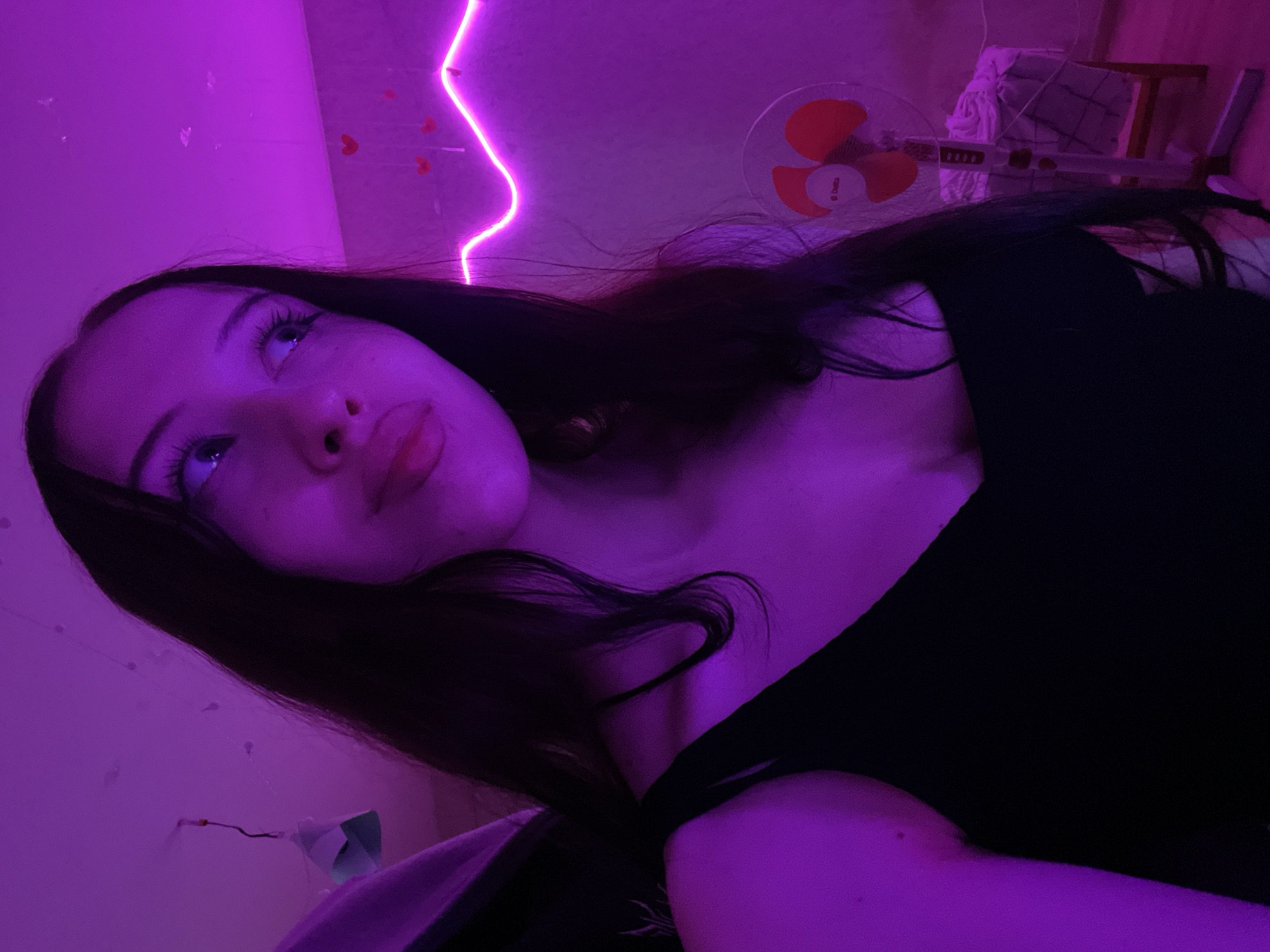 A young woman with long dark hair is posing for a selfie in a room illuminated by purple LED lights. She is wearing a black tank top. Her gaze is directed upwards and to the right. In the background, a pink neon light strip curves along the wall, adorned with small red hearts. To the right, a white oscillating fan stands on a small wooden stool next to a draped object. The setting appears to be a bedroom or personal space in Votkinsk, Russia.