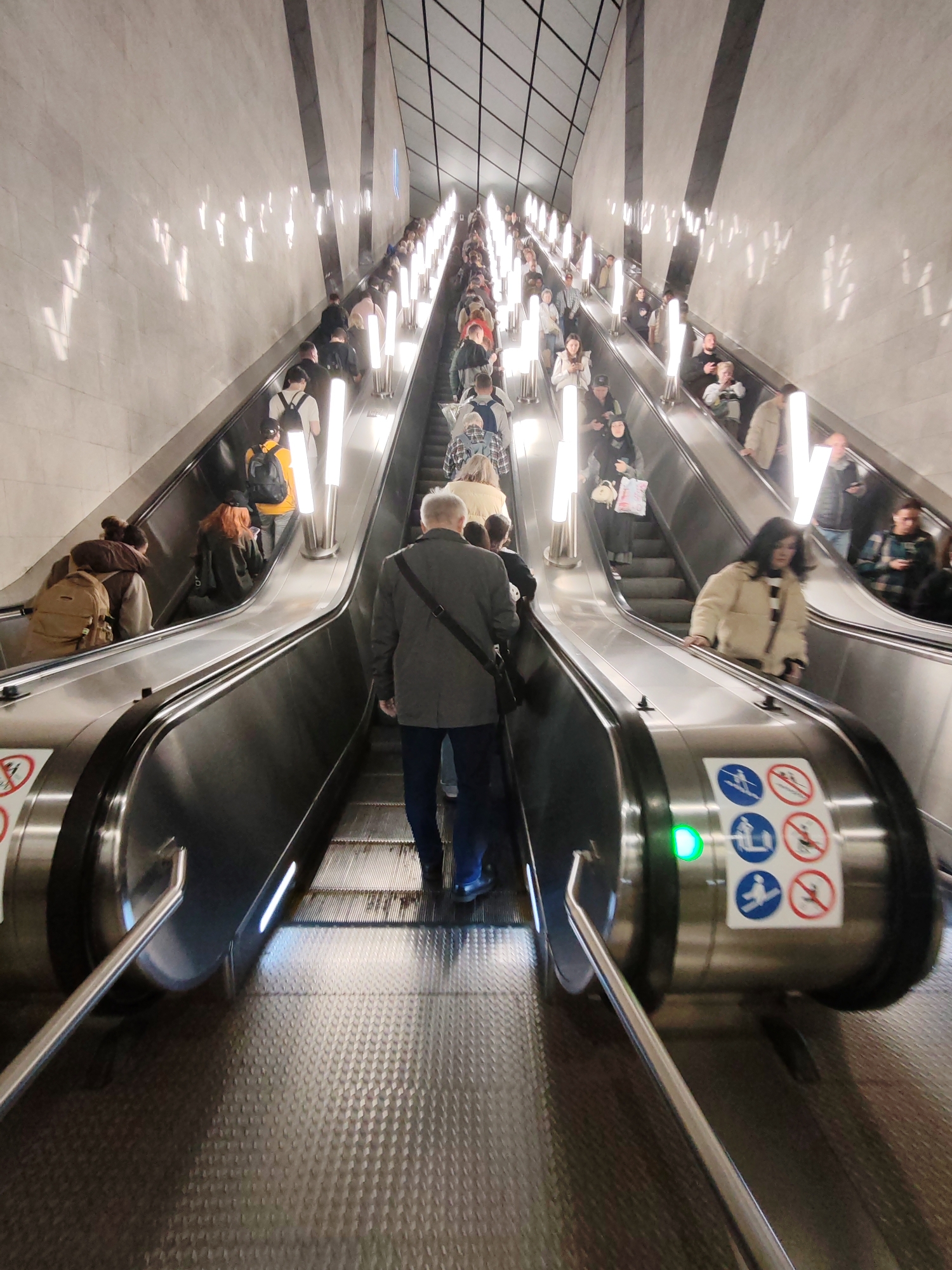 The image shows a wide escalator in what appears to be a modern subway station or large public transit hub. Numerous people are ascending and descending the escalators, many carrying backpacks and bags, suggesting a busy travel environment. The walls of the station are lined with vertical fluorescent lights, creating a bright, somewhat reflective surface. The escalators themselves are metallic with textured steps. Safety signs are visible on the side of the escalator, including icons for standing, not running, and no pets. A green indicator light is also visible on one of the escalators. The perspective is from someone on the escalator looking up and down.