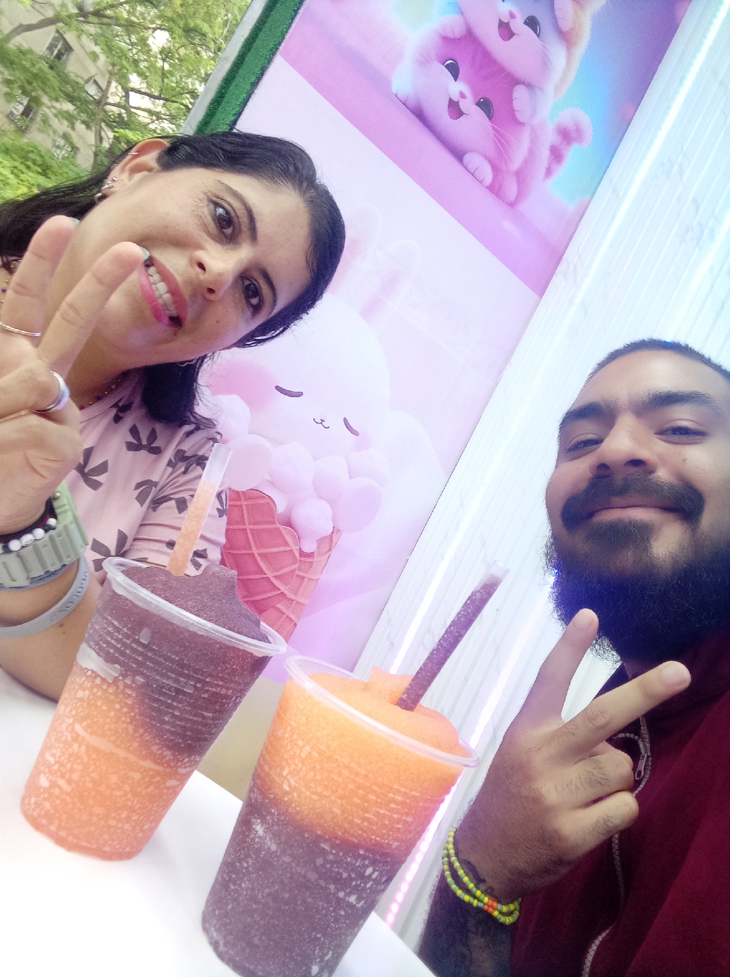 The image is a selfie of a woman and a man in front of a backdrop with cartoon animals and an ice cream cone. The woman, on the left, has dark hair, fair skin, and is wearing a patterned pink shirt. She is holding up a peace sign and smiling. The man, on the right, has a dark beard, mustache, and dark hair pulled back in a bun. He is wearing a maroon shirt, also holding up a peace sign, and smiling. In front of them, on a white surface, are two cups filled with layered frozen beverages, one with dark and orange layers, and the other with orange and dark layers. Each cup has a straw. The backdrop behind them features images of cartoon cats and a smiling cloud shaped like an ice cream cone. The scene is brightly lit, and a glimpse of green foliage is visible at the top left corner of the frame. The location is Guarenas, Venezuela.