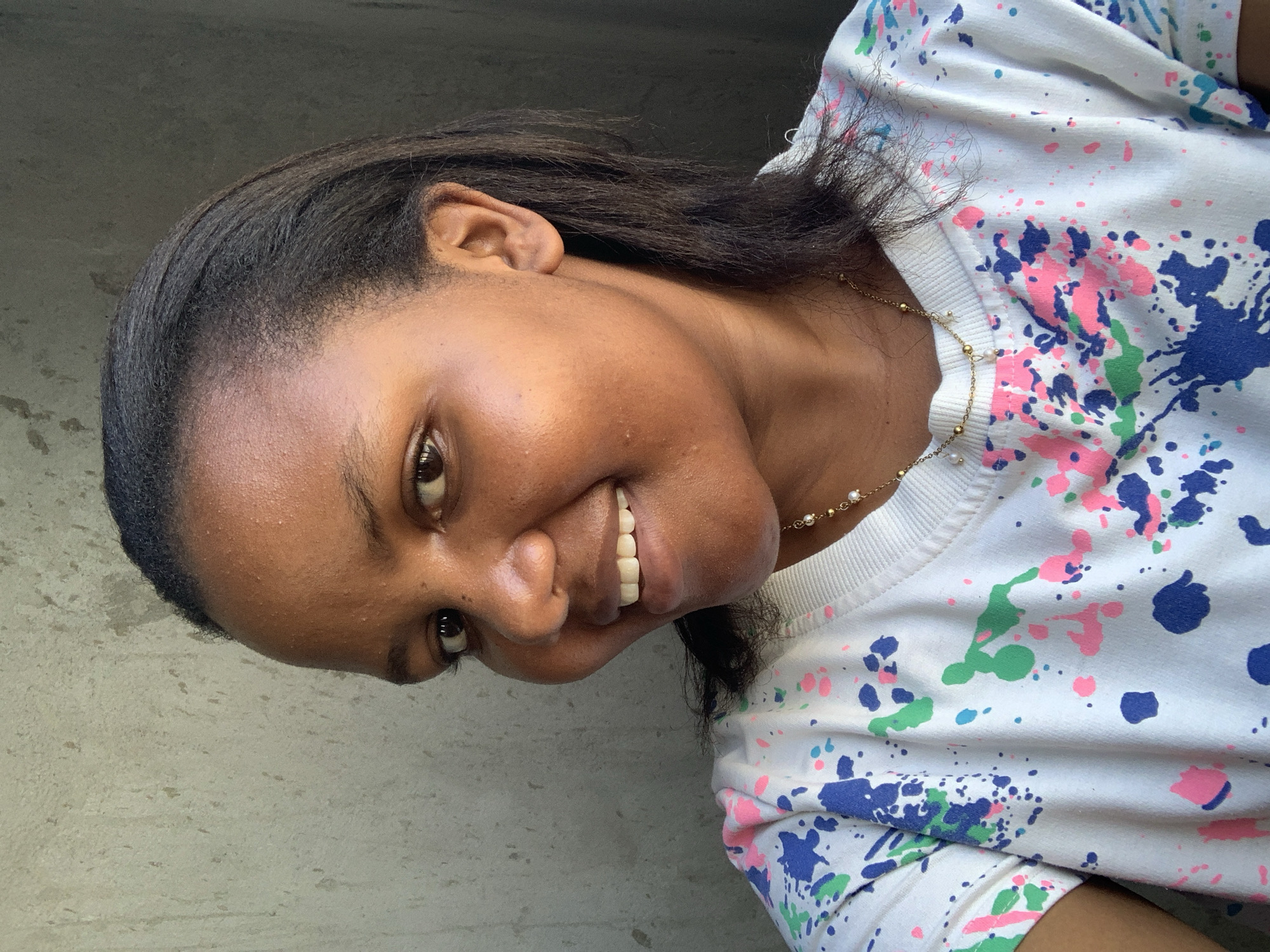 A young woman smiles at the camera, wearing a white shirt with splashes of pink, blue, and green paint. She is wearing a gold necklace. The background appears to be a gray wall. It is impossible to identify the location in Dar es-Salaam, Tanzania from the image provided.
