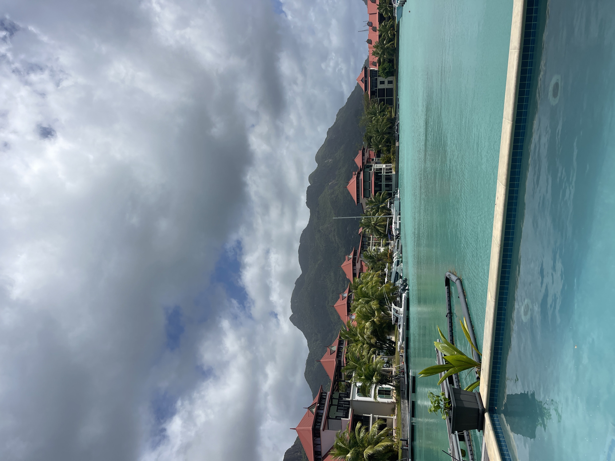 The image depicts a waterfront scene in Providence, Seychelles. In the foreground, a portion of a swimming pool is visible, with its blue tiled edge leading into the clear turquoise water of a harbor. Beyond the pool, a pier with dark planks extends into the harbor. A black planter with two green plants sits on the pier.
The harbor is filled with several white boats moored along the docks. In the middle ground, a row of buildings with distinctive red, pyramidal roofs and white facades lines the waterfront, interspersed with lush green palm trees. These buildings appear to be part of a resort or residential complex.
In the background, a range of green, mountainous terrain rises above the buildings. The sky is partially overcast with large, gray clouds, suggesting a mixed weather day. The overall impression is one of a tranquil, tropical resort environment.