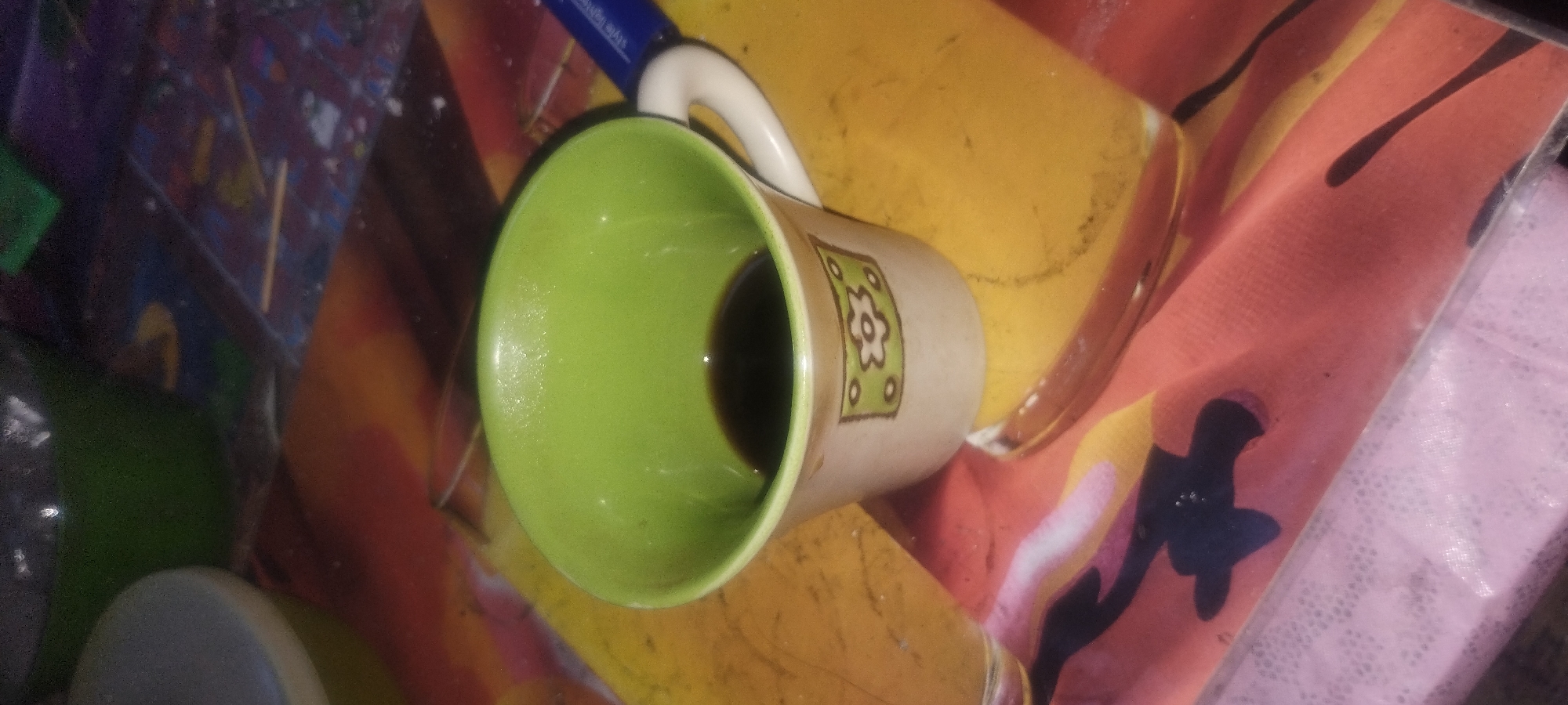 A close-up, high-angle shot captures a white ceramic mug with a green interior, filled with dark liquid, likely coffee. The mug sits on a colorful, patterned surface that appears to be a placemat or tablecloth. To the right of the mug, a blue pen with white lettering is visible, lying on the same surface. The background is out of focus but suggests a cluttered environment, with hints of other objects and a blurred green bowl in the upper left. The overall lighting is dim, creating a slight grainy effect. The image is not geotagged and does not contain any information about its location in Zaporizhzhia, Ukraine.
