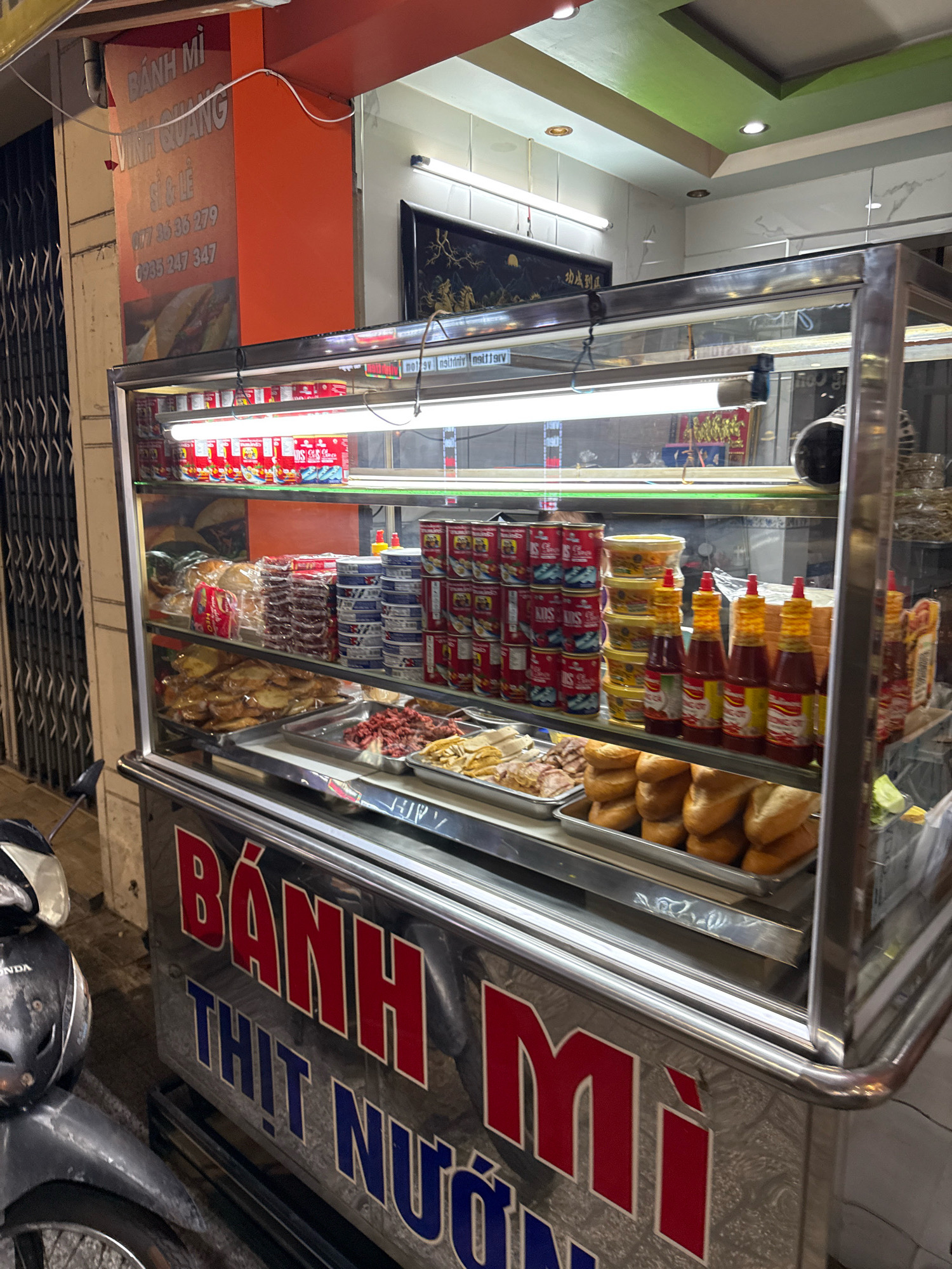 The image shows a food stall on a street in Nha Trang, Vietnam, specializing in Banh Mi, a Vietnamese baguette sandwich. The stall is illuminated by fluorescent lights and features a glass display case filled with various ingredients for the sandwiches. On the top shelf, rows of canned goods and what appear to be butter or spreads are visible. The middle shelf holds plastic-wrapped loaves of bread and processed meats. The bottom shelf displays cooked meats, sliced bread, and various condiments, including several bottles of chili sauce. The stall's signage, prominently displayed on the front, reads "BÁNH MÌ THỊT NƯỚNG" in large red and blue letters, indicating the type of food sold. A portion of a motorcycle is visible in the lower left corner of the frame. The overall scene suggests a bustling street food vendor operating in an urban environment at night.