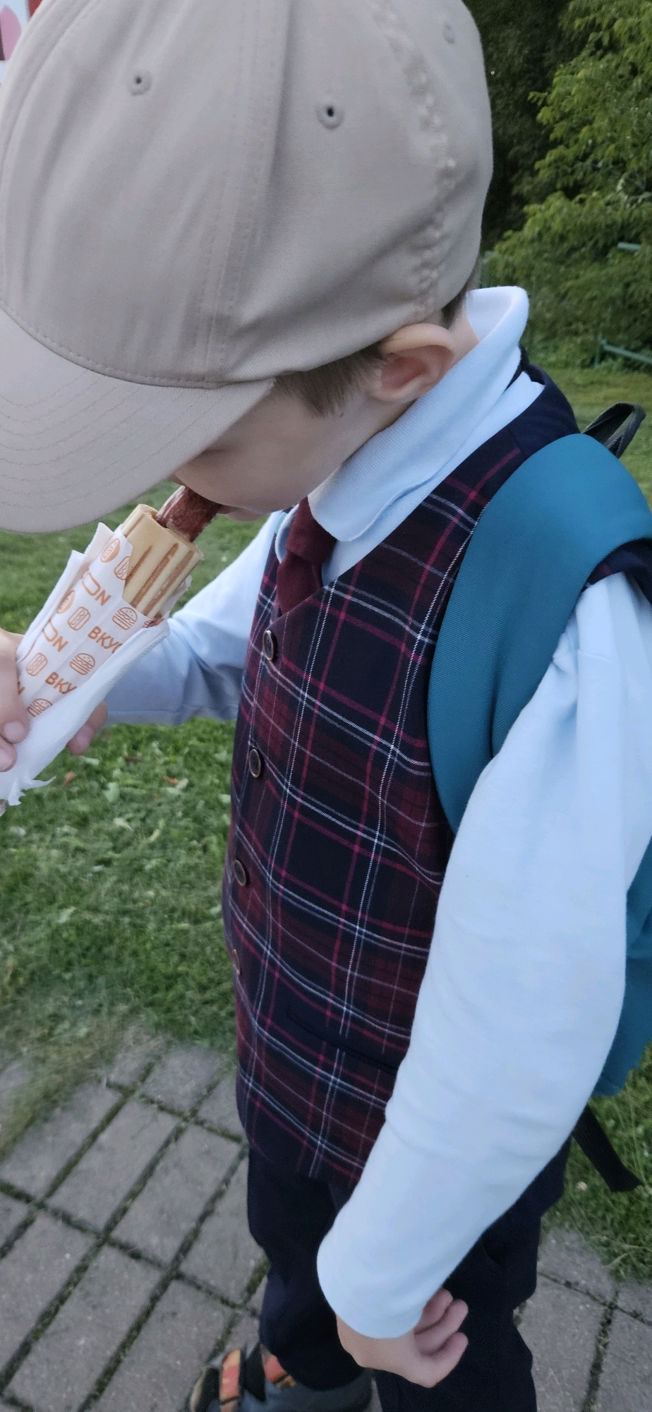 A child wearing a light-colored baseball cap, a light blue collared shirt, a dark red and blue plaid vest, and a dark backpack is holding and eating a hot dog wrapped in a paper sleeve. The child is outdoors with a grassy area and a paved path visible. The hot dog wrapper has a pattern with images of hot dogs, hamburgers, and text that appears to be in Cyrillic. The child's face is partially obscured by the cap and the hot dog. The scene is likely a casual outdoor setting.