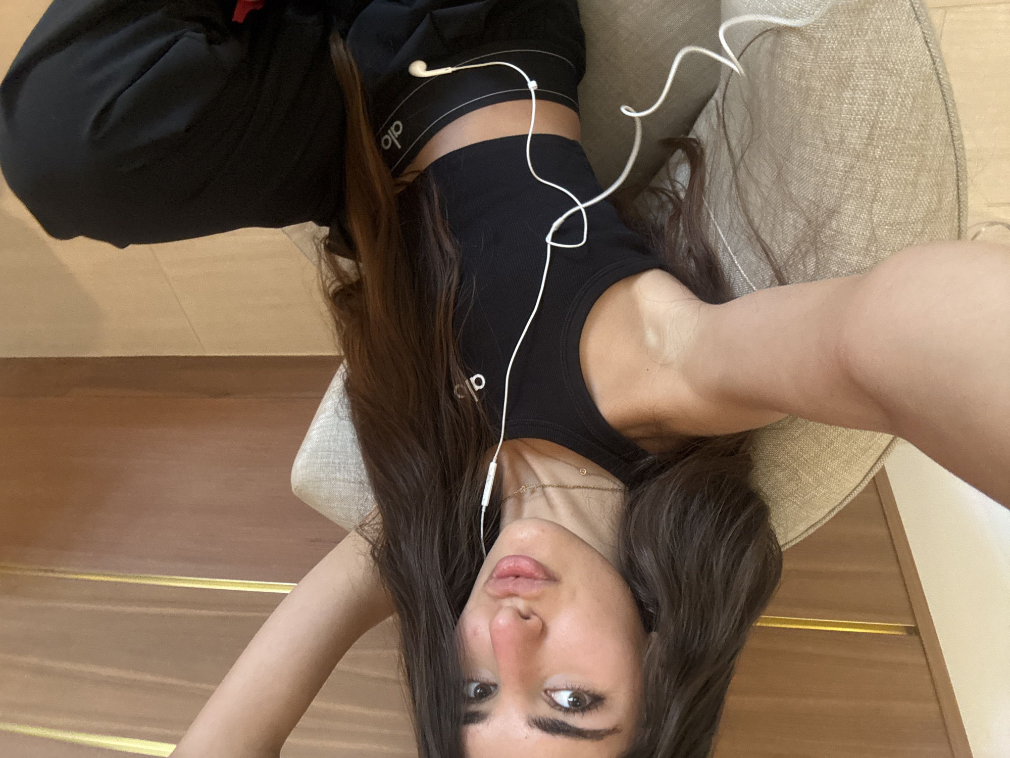 A young woman with long, dark hair is sitting in a chair, holding a phone and taking a selfie. She is wearing a black tank top and black pants, both with the "alo" logo visible. She has white earbuds in her ears. The background appears to be an indoor setting with wooden paneling and a textured chair. The lighting suggests it is daytime.