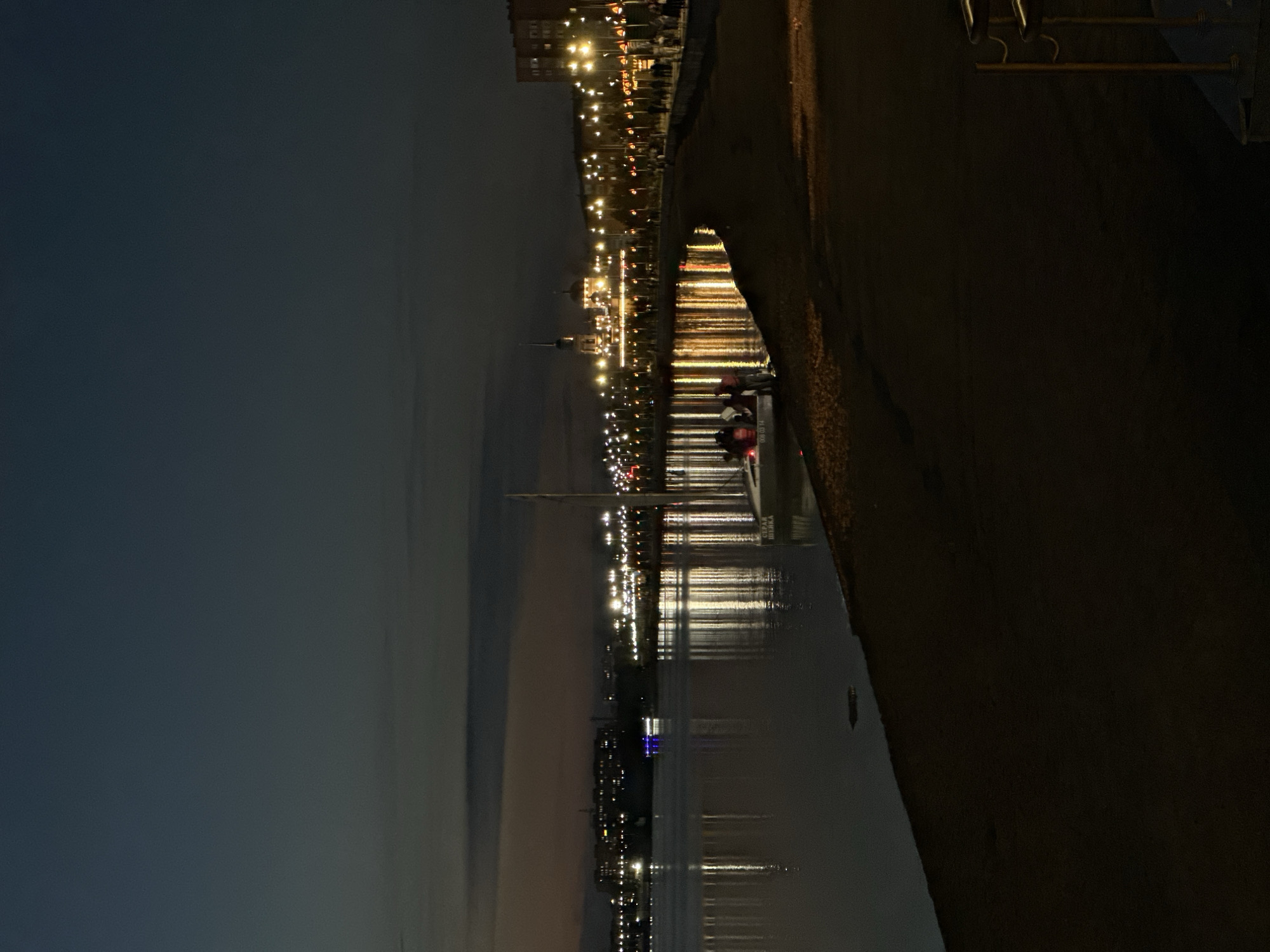 The image depicts a nighttime scene along a waterfront in Kokshetau, Kazakhstan. The sky is dark blue with subtle hints of lighter hues near the horizon, indicating twilight or a cloud-obscured night. A calm body of water occupies the left side of the frame, reflecting the lights from the city on the opposite bank. These lights are varied, with some appearing as bright white and yellow streaks, while others are more diffused. A boat with illuminated windows is visible on the water in the middle ground.
The right side of the image shows a sandy embankment leading to a promenade or walkway lined with streetlights. Several people are present on the promenade, appearing as small figures walking or standing. A few individuals are also gathered near a boat docked at the edge of the water on the left side of the embankment. In the far background, city buildings and structures are illuminated, creating a sense of urban presence. A sailboat mast is visible in the foreground on the right, and a section of a metal railing is also present. The overall atmosphere is tranquil, with the artificial lights providing the primary illumination against the dark natural surroundings.