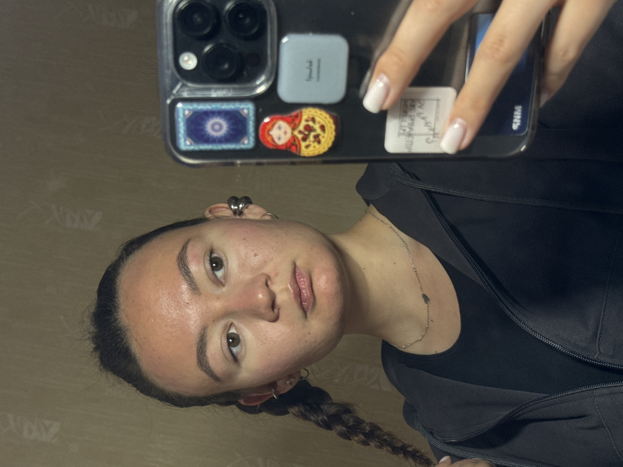 A fair-skinned person with light brown eyes and dark brown hair in a braid is taking a selfie with a smartphone. The person is wearing a black top and a dark jacket, along with a silver necklace. The smartphone case has stickers of a matryoshka doll, a blue and white decorative tile, and other opaque rectangular stickers. The background is a plain beige wall. The location details (Chaikovsky, Russia) cannot be verified from the image.