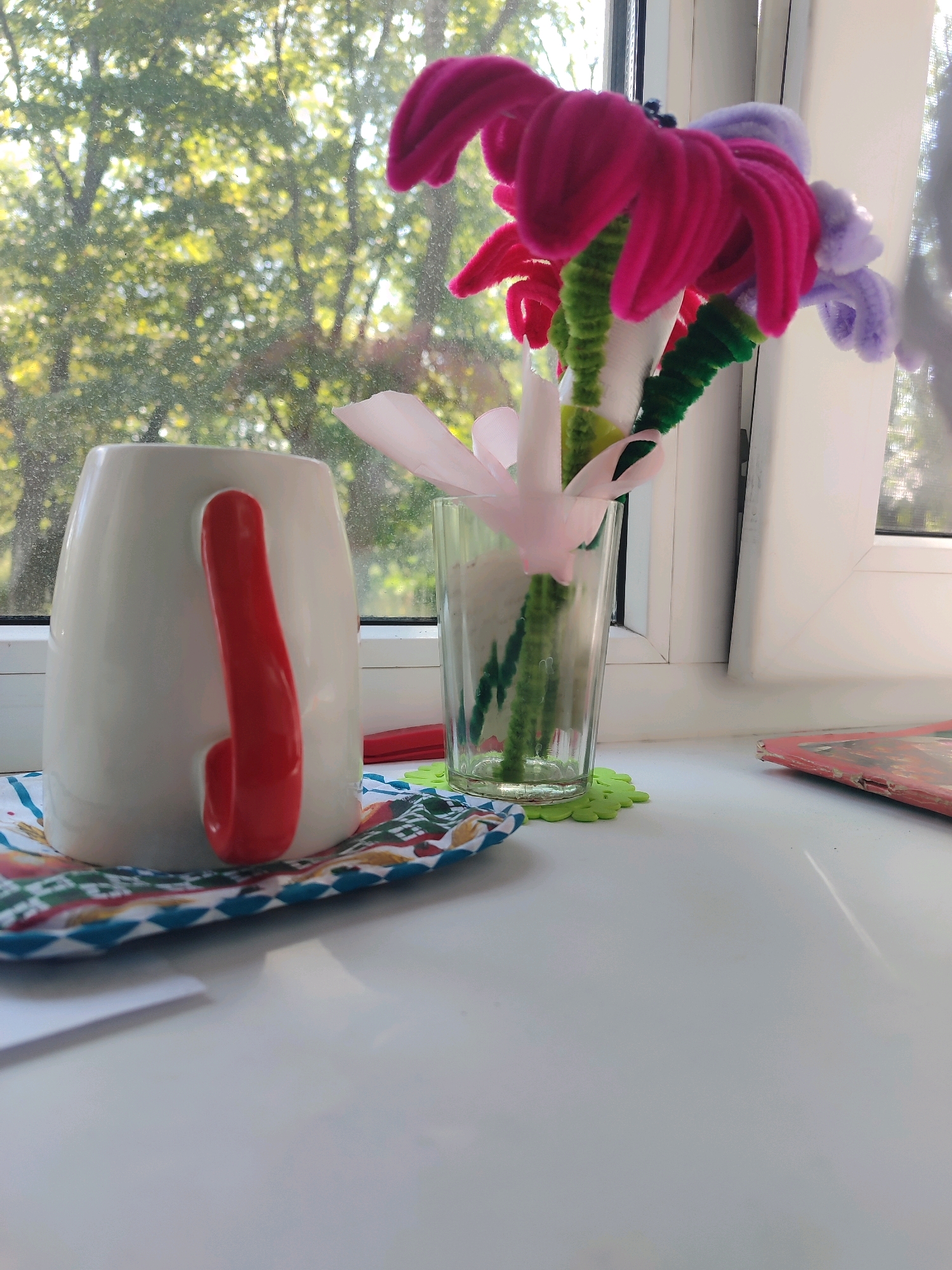 An indoor still life photograph is captured from a slightly low angle, focusing on a white ceramic mug with a bright red handle. The mug is positioned on a colorful patterned coaster, which sits on a white ledge, likely a windowsill. Behind the mug, a glass vase holds a bouquet of pipe cleaner flowers in shades of fuchsia, pink, and pale purple, with green pipe cleaner stems. The flowers appear to be handmade. To the right of the vase, part of a white window frame is visible, with dappled sunlight streaming through what appears to be foliage outside, suggesting the scene is illuminated by natural light. The background shows the blurred shapes of trees and leaves, indicating an outdoor view. On the far right of the ledge, a portion of a red-covered book or magazine is visible. The overall impression is a domestic setting, possibly a windowsill in a home. The exact location within a city is not discernible from the image.