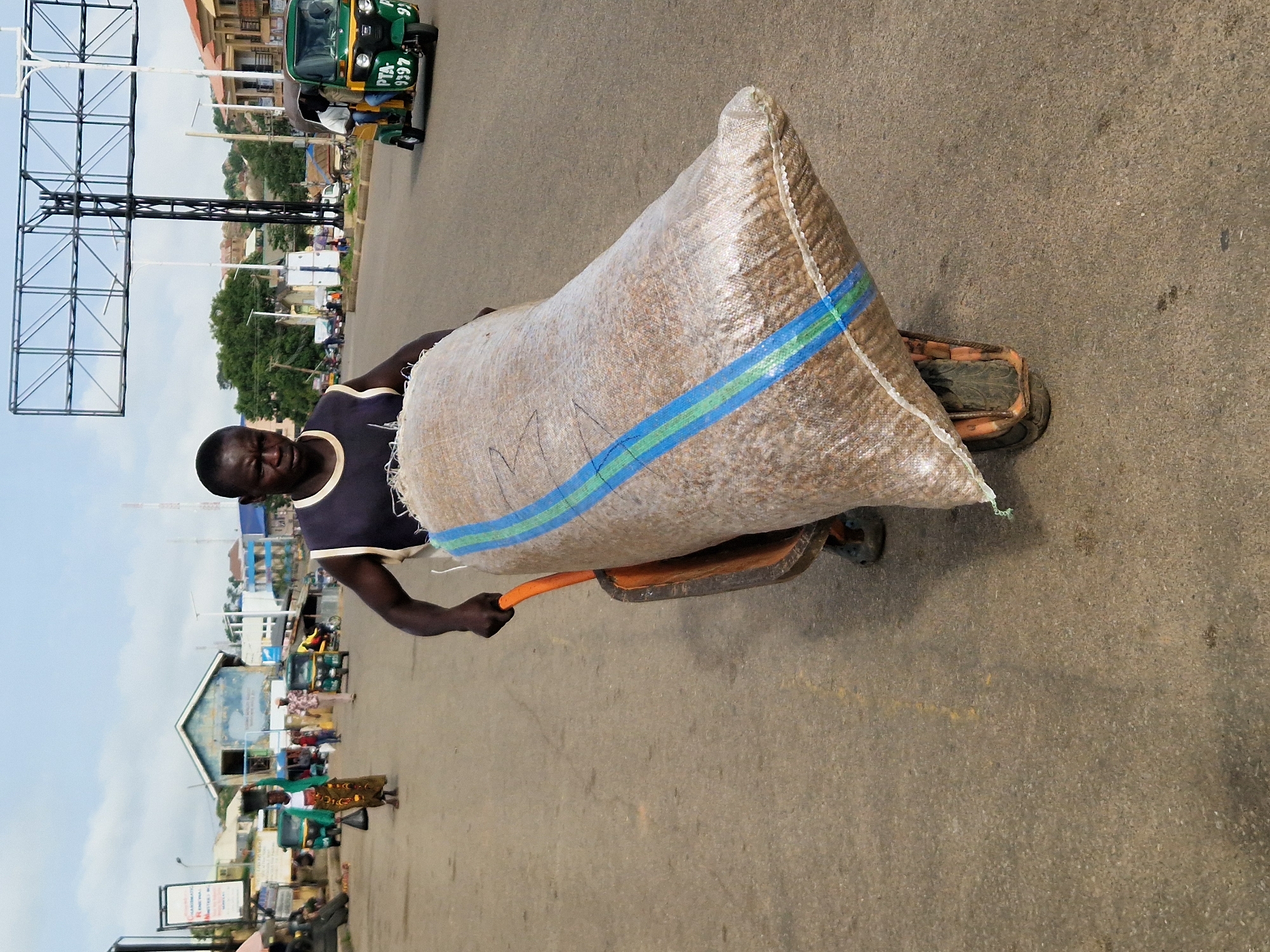 A medium shot shows a man in Kabong, Nigeria, pushing a wheelbarrow filled with a large sack of grain. The man is wearing a dark blue tank top and dark shorts, and he is focused on his task. The sack is made of rough, woven material and has blue and green stripes running horizontally across it. The wheelbarrow has an orange handle and a single wheel with a black tire. The man is on a paved road, with buildings and other vehicles visible in the background. A green and yellow tuk-tuk is driving away from the viewer on the right side of the image. The sky is overcast with a few clouds. The scene appears to be a busy market or street in a city.