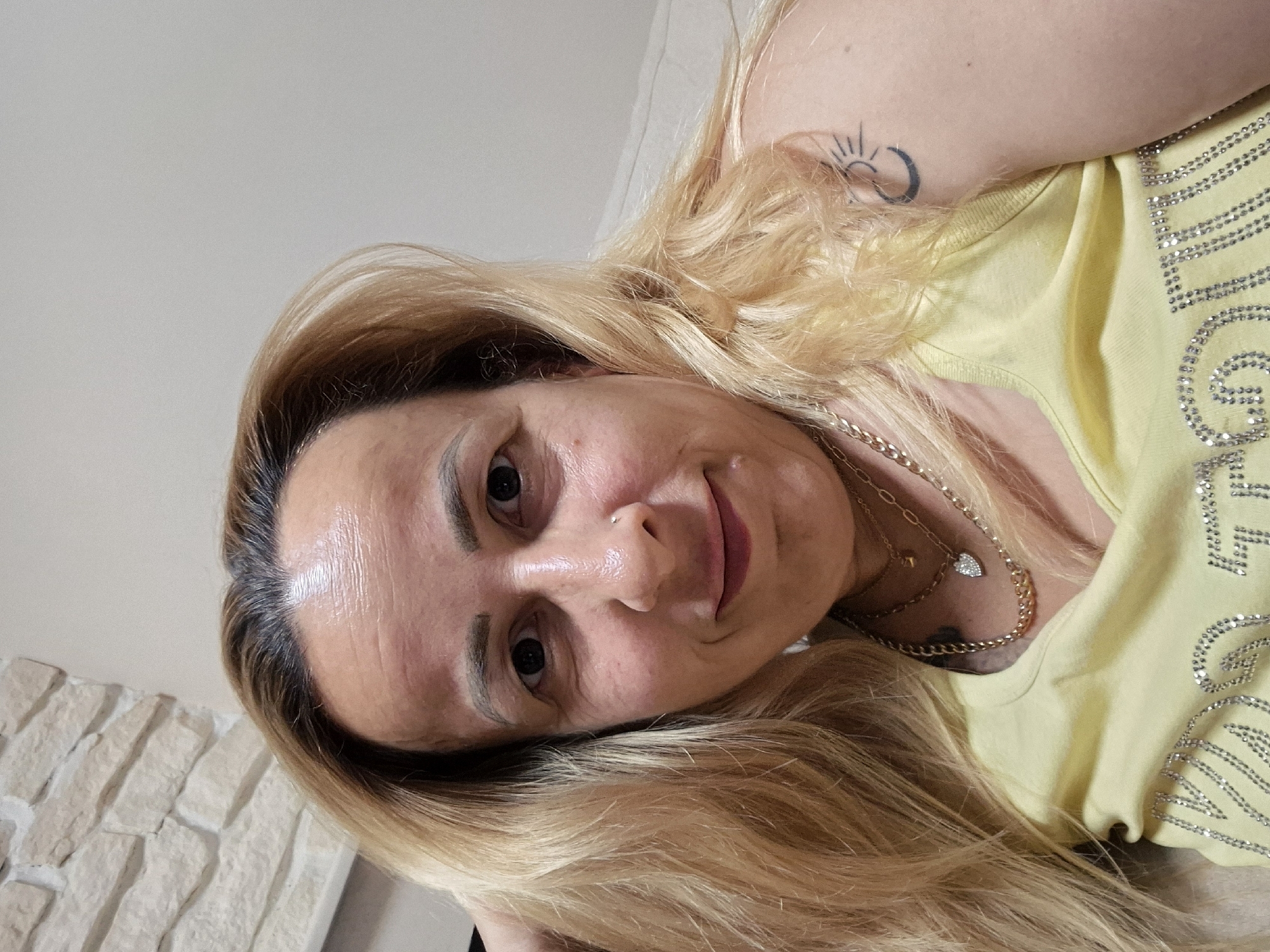 A medium shot shows a light-skinned person with blonde hair and a fair complexion, looking directly at the camera. Their hair is parted on the right and falls past their shoulders. The person is wearing a light yellow tank top adorned with rhinestones on the chest. A gold necklace with a heart pendant and a cuban link chain is visible around their neck. A small nose ring is present on their left nostril. A tattoo of a crescent moon with a sun inside is visible on their right upper arm. The background consists of a stone wall on the left and a plain light-colored wall on the right. The person appears to be indoors, possibly in a living room.