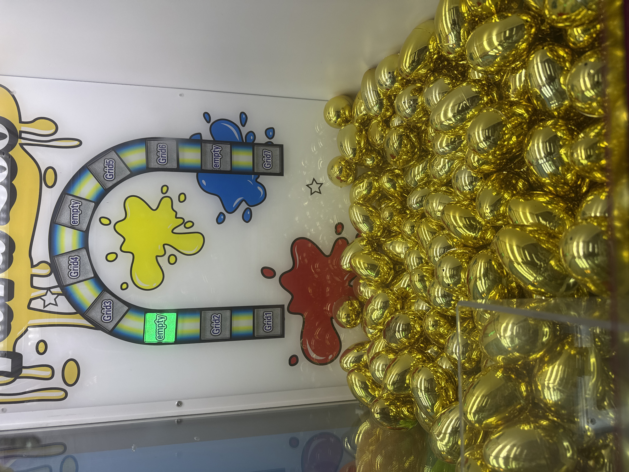An indoor scene in Sliven, Bulgaria, showcases a section of what appears to be an arcade game or prize vending machine. The right side of the image is dominated by a large pile of numerous shiny, reflective, gold-colored, egg-shaped capsules or balls, suggesting they contain prizes or are game tokens. They are densely packed behind a clear plastic barrier, reflecting distorted light and shapes from the surroundings.
On the left side of the image, part of the game's mechanism is visible, featuring a white background adorned with stylized yellow, red, and blue paint splatters. A U-shaped track, outlined with alternating yellow and blue stripes, curves across the panel. This track contains several rectangular slots. Visible text labels on these slots include "Grid1", "Grid2", "Grid3", "Grid4", "Grid5", "Grid6", "Grid7", and multiple instances of "empty". One of the "empty" slots, positioned roughly in the center of the visible track, is brightly illuminated with a vibrant green light, indicating a current state or target within the game. The arrangement suggests a game where the golden capsules might traverse this track, landing in designated "Grid" or "empty" slots as part of gameplay. No people are visible, and the time of day is not discernible, but the bright, clean aesthetic points to a well-maintained entertainment venue.