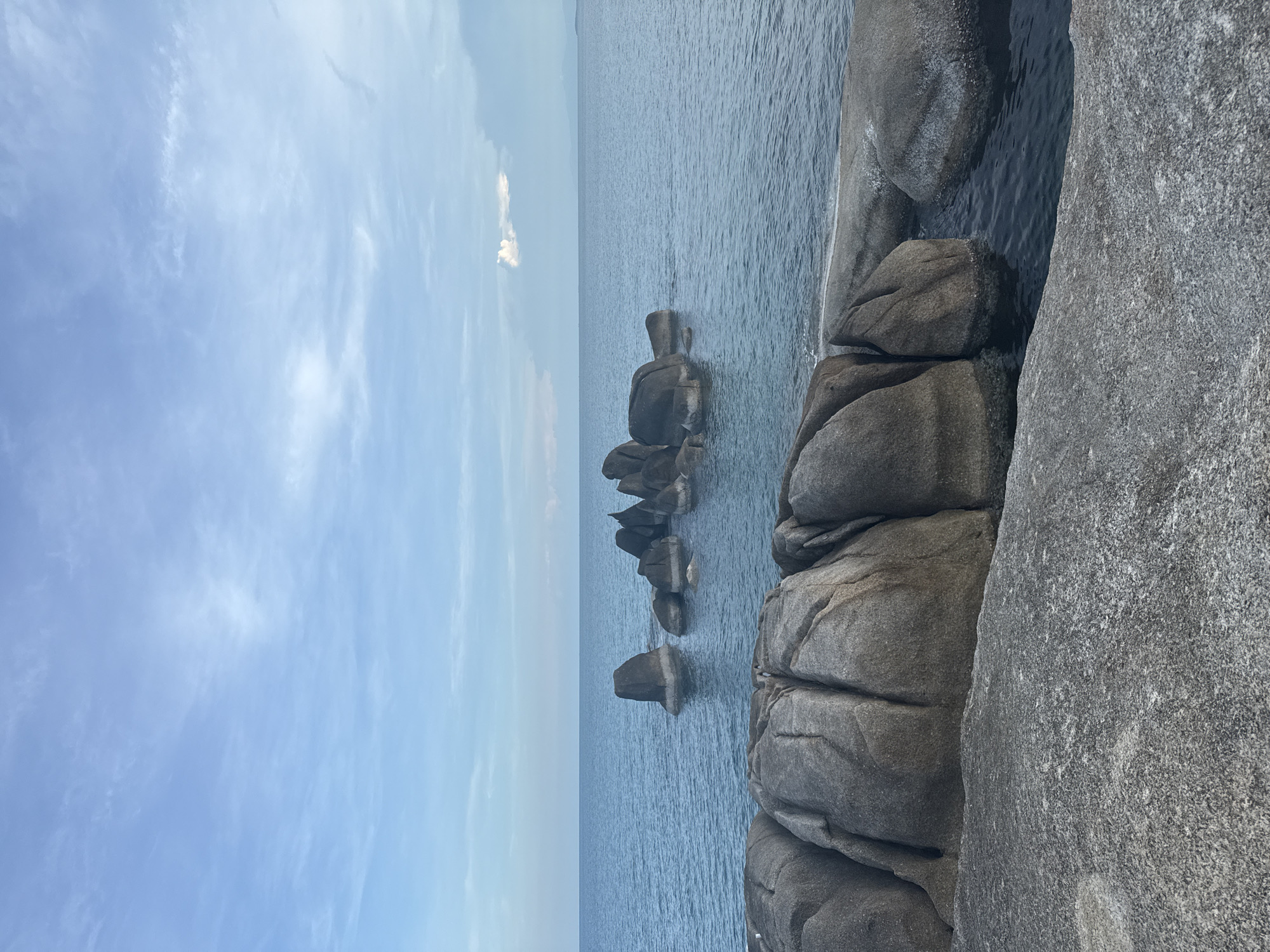 The image depicts a serene seascape with large, rounded granite boulders scattered across the foreground and jutting out from the calm, blue ocean in the middle ground. The foreground is dominated by smooth, weathered rocks, some partially submerged in clear water. In the distance, a cluster of similarly shaped boulders forms a small islet. The sky above is a soft blue with wispy white clouds, suggesting it is either early morning or late afternoon. The overall scene evokes a sense of tranquility and natural beauty. The location is identified as Baan Hua Thanon, Thailand, a coastal village known for its picturesque beaches and rock formations. No people or animals are visible in the image.
