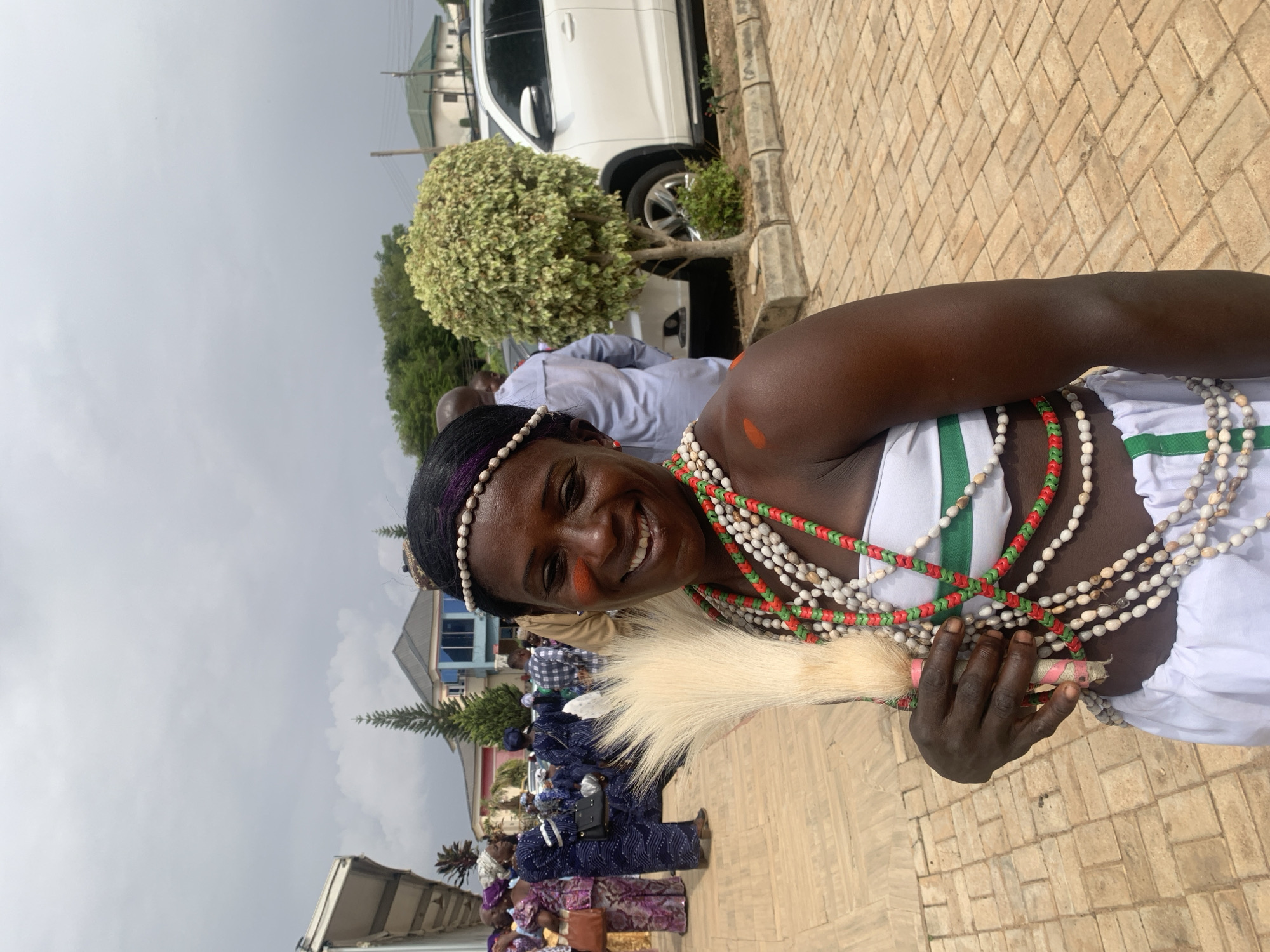 An outdoor shot in Jos, Nigeria, captures a dark-skinned woman smiling at the camera. She wears a white dress with green stripes and an array of beaded necklaces in white, red, and green. Her hair is adorned with a beaded headband, and she has orange face paint on her cheek. She holds a white fluffy item, possibly a ceremonial object. In the background, a crowd of people is visible, along with a white car, trees, and buildings. The ground is paved with light brown bricks. The sky is overcast.