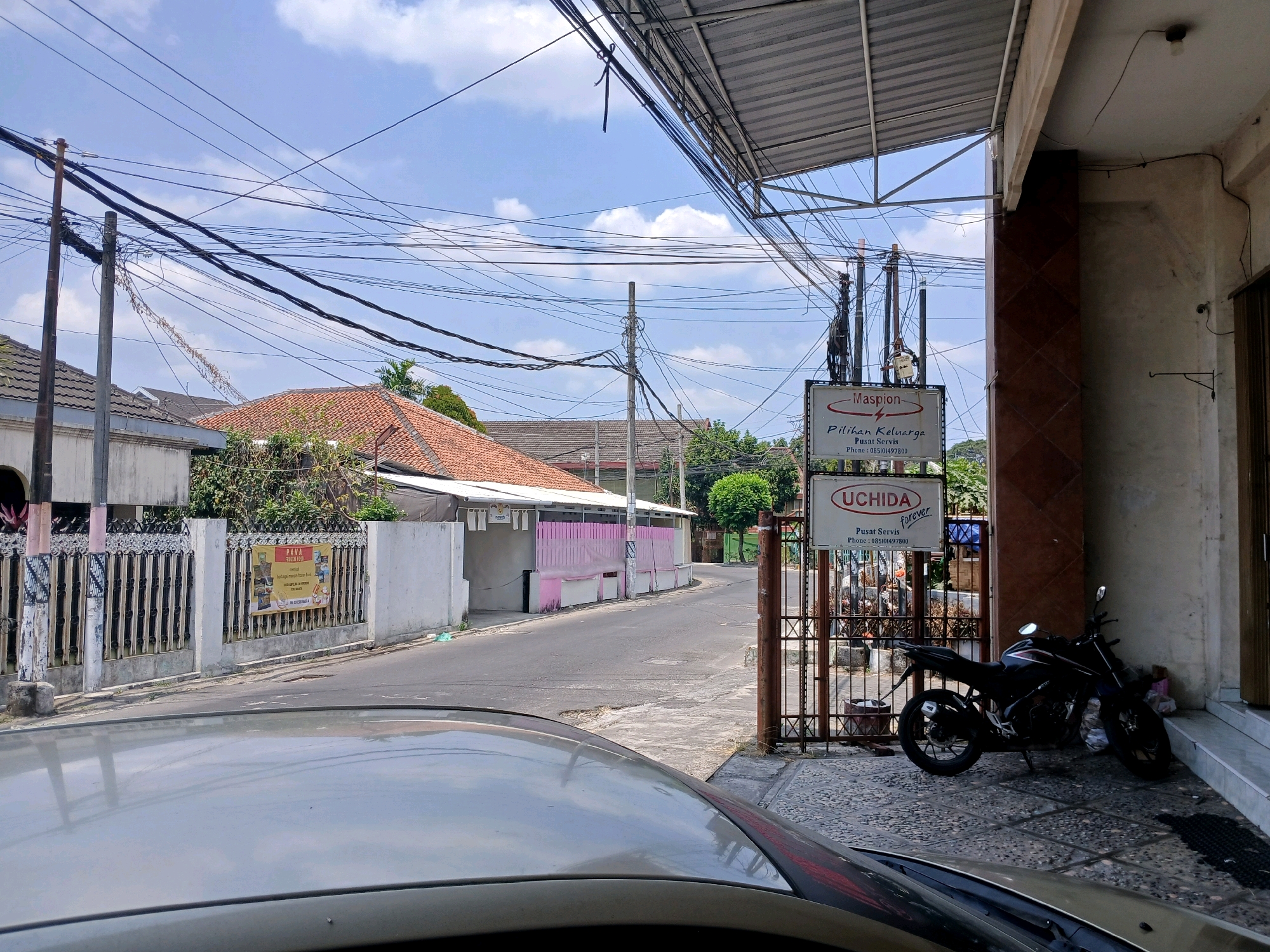 A motorcycle is parked in front of a building with a sign that reads "UCHIDA forever." The building appears to be a business, possibly a service center, as the sign also includes "Pusat Servis" (Service Center) and a phone number. The motorcycle is black and is parked on a paved area.
In the background, there is a street with houses on either side. The houses have tiled roofs and white walls. There are many electrical wires strung across the sky, typical of an urban environment. A fence with a banner is visible on the left side of the street. The sky is blue with some clouds. The overall impression is a street scene in an Indonesian city, likely during the daytime, given the lighting. The presence of the "UCHIDA forever" sign and the style of the houses suggest this could be Depok, Indonesia, given the commonality of such businesses and residential architecture in the region.
