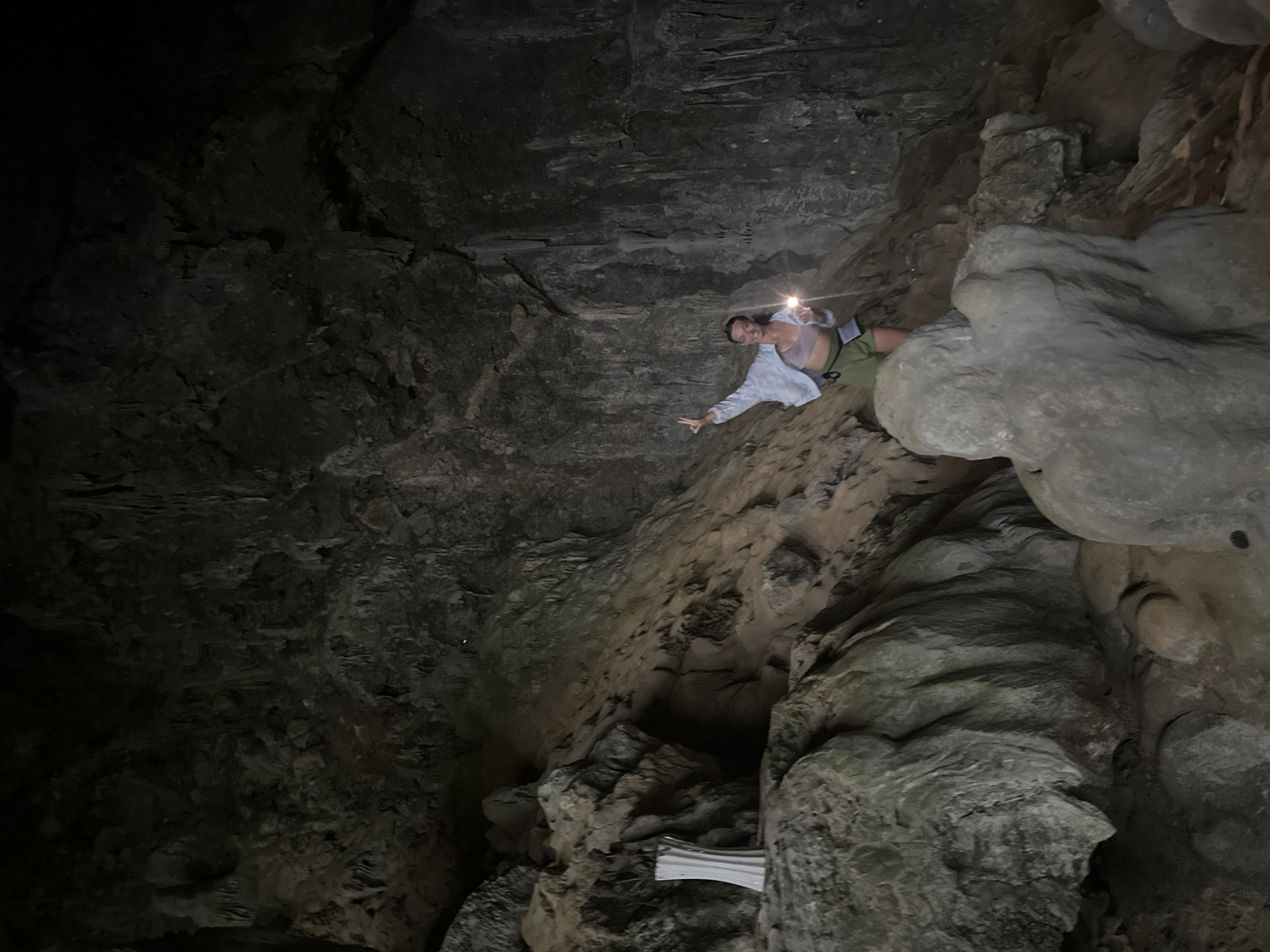 A young woman stands in a dimly lit cave, holding a flashlight and making a peace sign with her fingers. She is wearing a light-colored open jacket over a pink sports bra and olive green shorts. The cave walls are rough and rocky, with stalactites hanging from the ceiling. To the left of the woman, there is a white, vase-like object on a rocky ledge. The ground is uneven and covered with rocks and debris. The overall impression is one of exploration and adventure in a natural environment. The location is within a cave in Mai Chau, Vietnam.