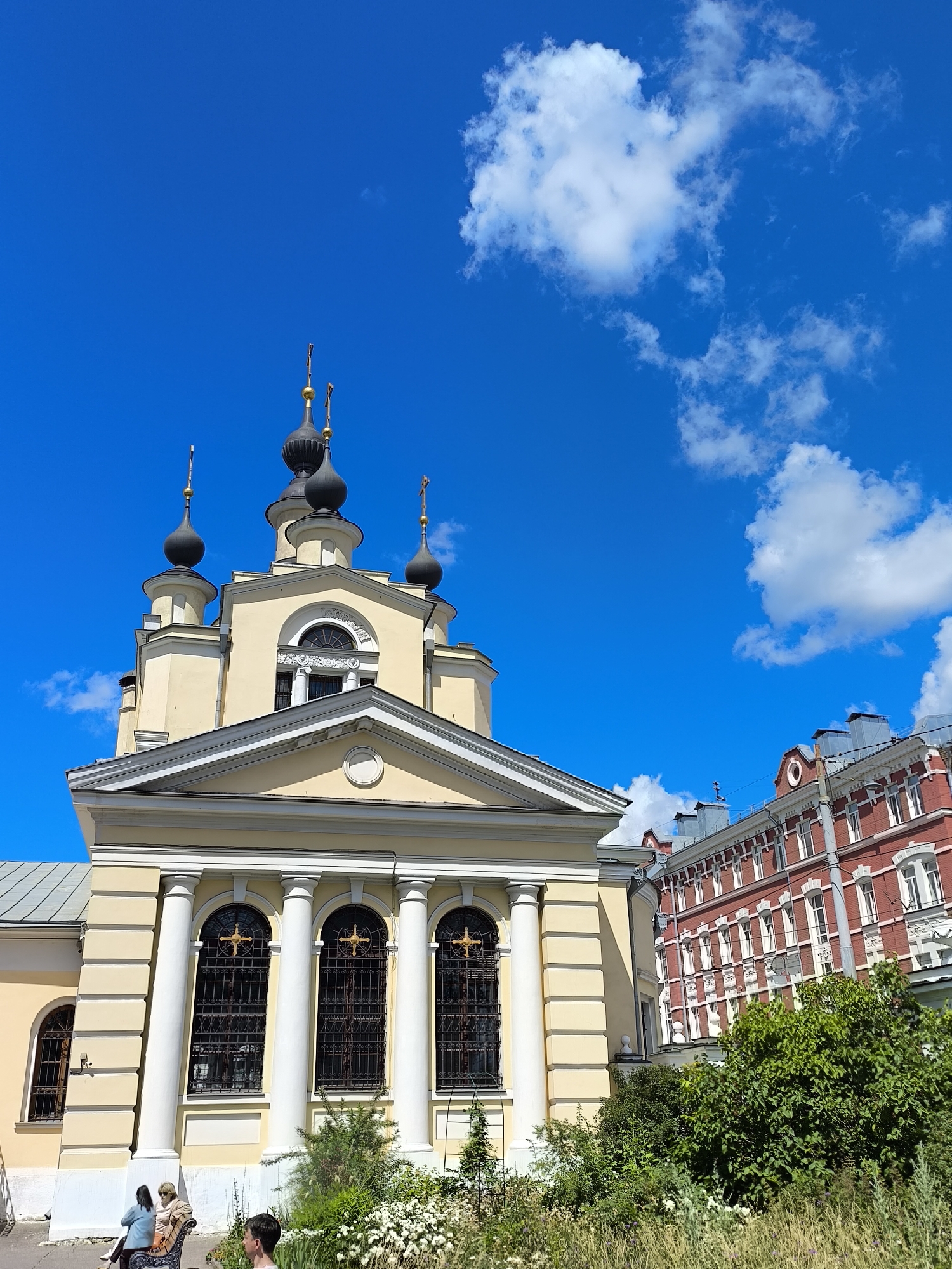 An exterior shot depicts a religious building with a pale yellow facade and several dark, onion-shaped domes topped with golden crosses. The building features white classical columns and arched windows with black metal grates. To the right, a red brick building with white trim and numerous windows is visible. Lush green bushes and tall, dry grass grow in the foreground. Two individuals are seated on a bench to the left, and another person stands to the right. The scene is set against a bright blue sky with scattered white clouds.